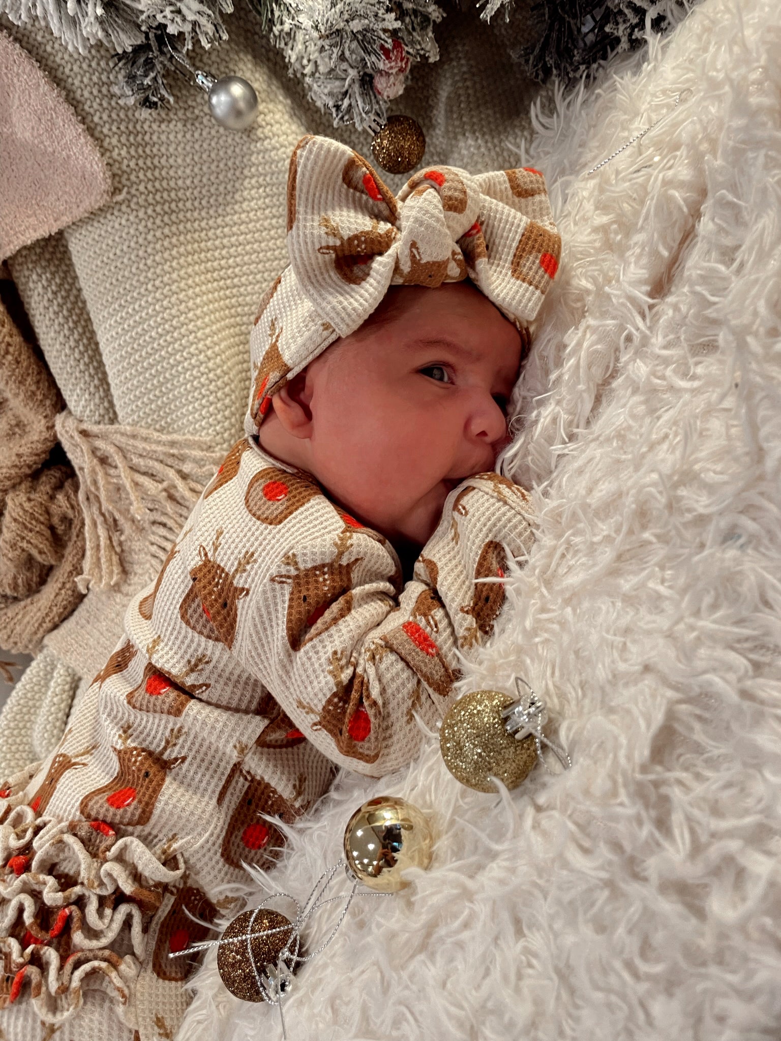 A baby in a reindeer-patterned outfit and bow rests on a soft blanket with festive ornaments nearby.
