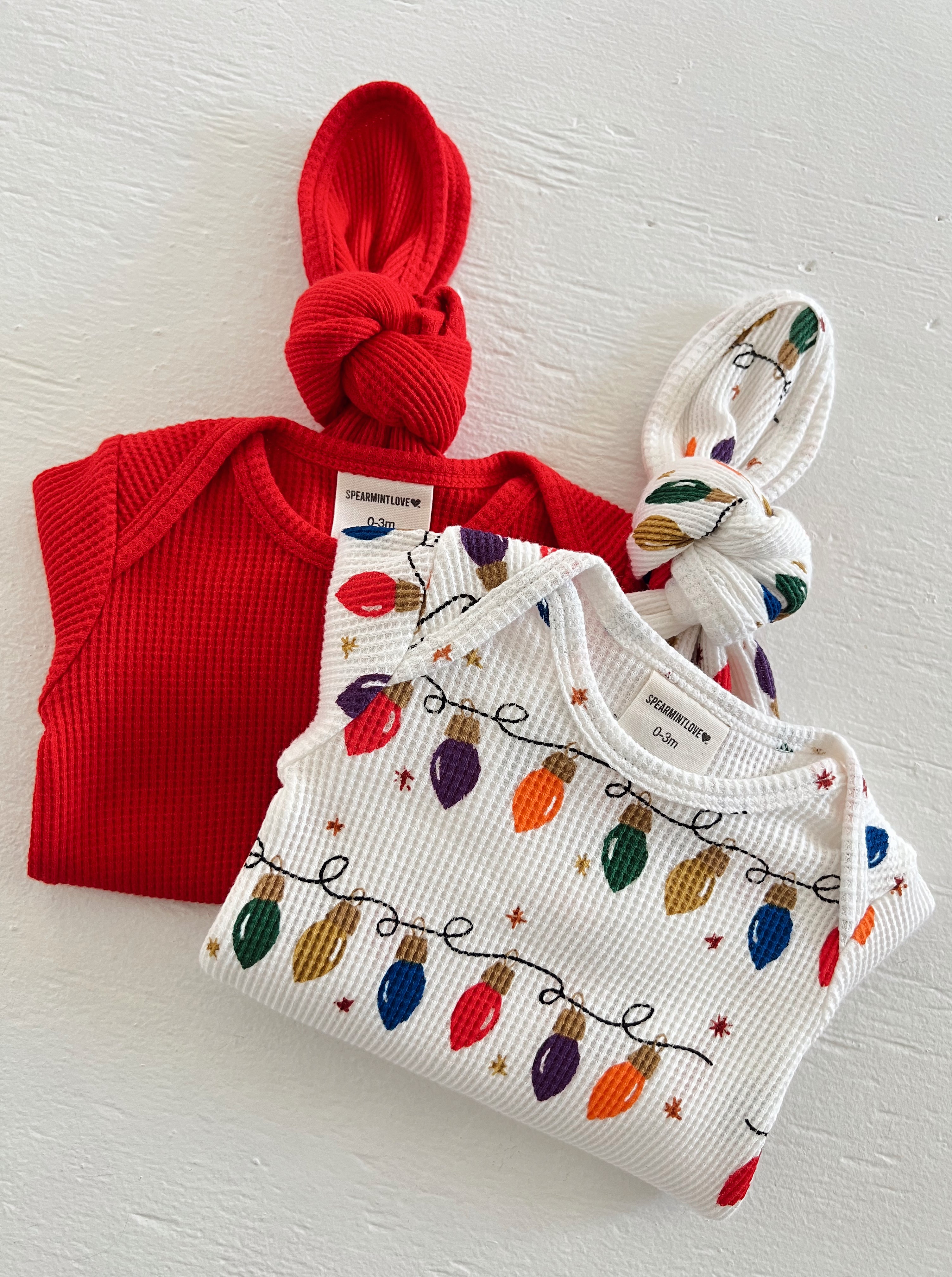 Red baby onesie and festive white outfit with colorful string lights, displayed on a neutral background.