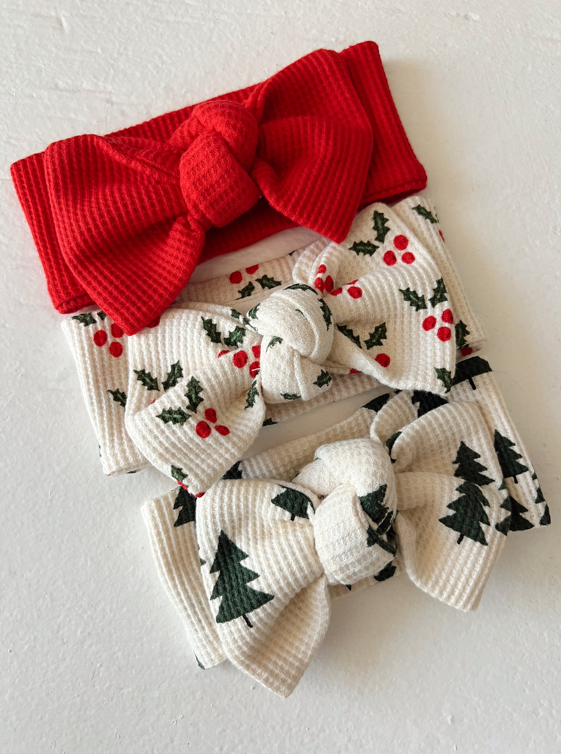 Three festive headbands: a red one, and two with holly and evergreen tree prints. Perfect for holiday styling.
