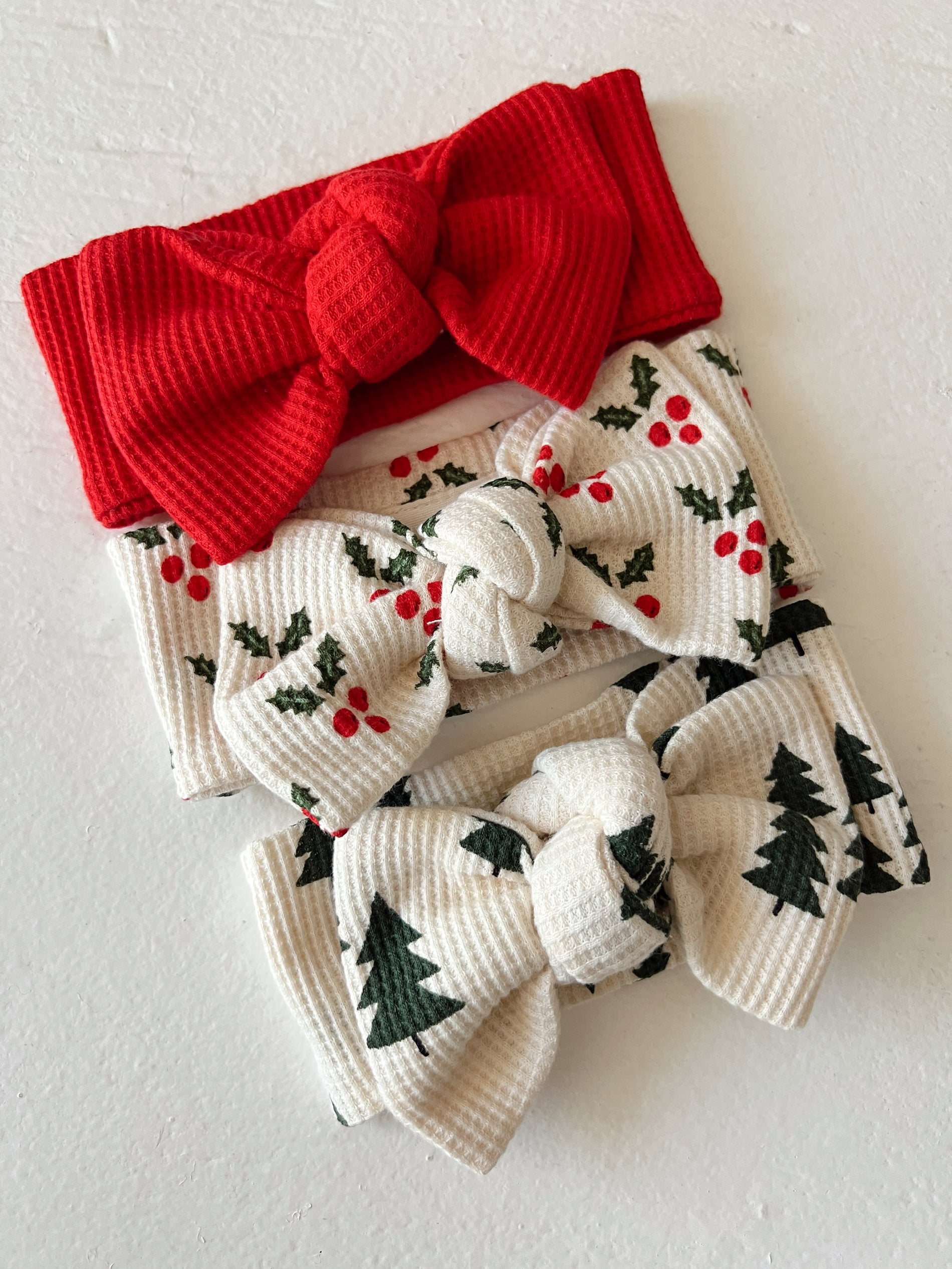 Three festive headbands: a red one, and two with holly and evergreen tree prints. Perfect for holiday styling.