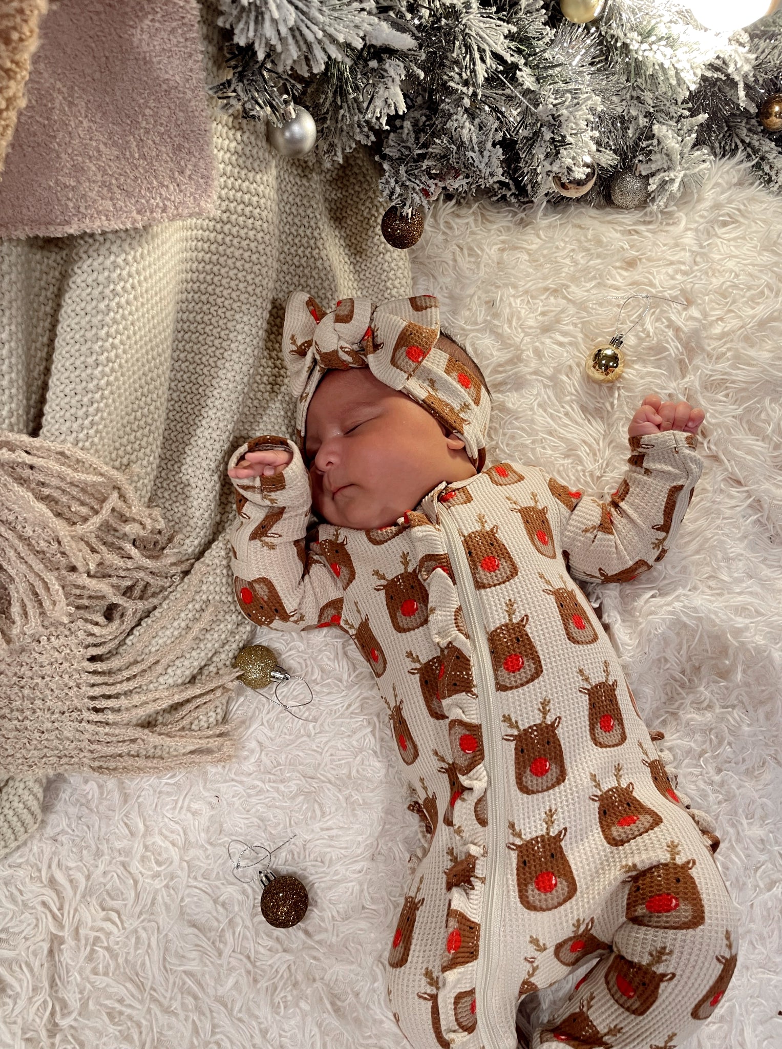 Infant in a festive reindeer onesie sleeping on a cozy blanket with holiday decorations and soft lighting.