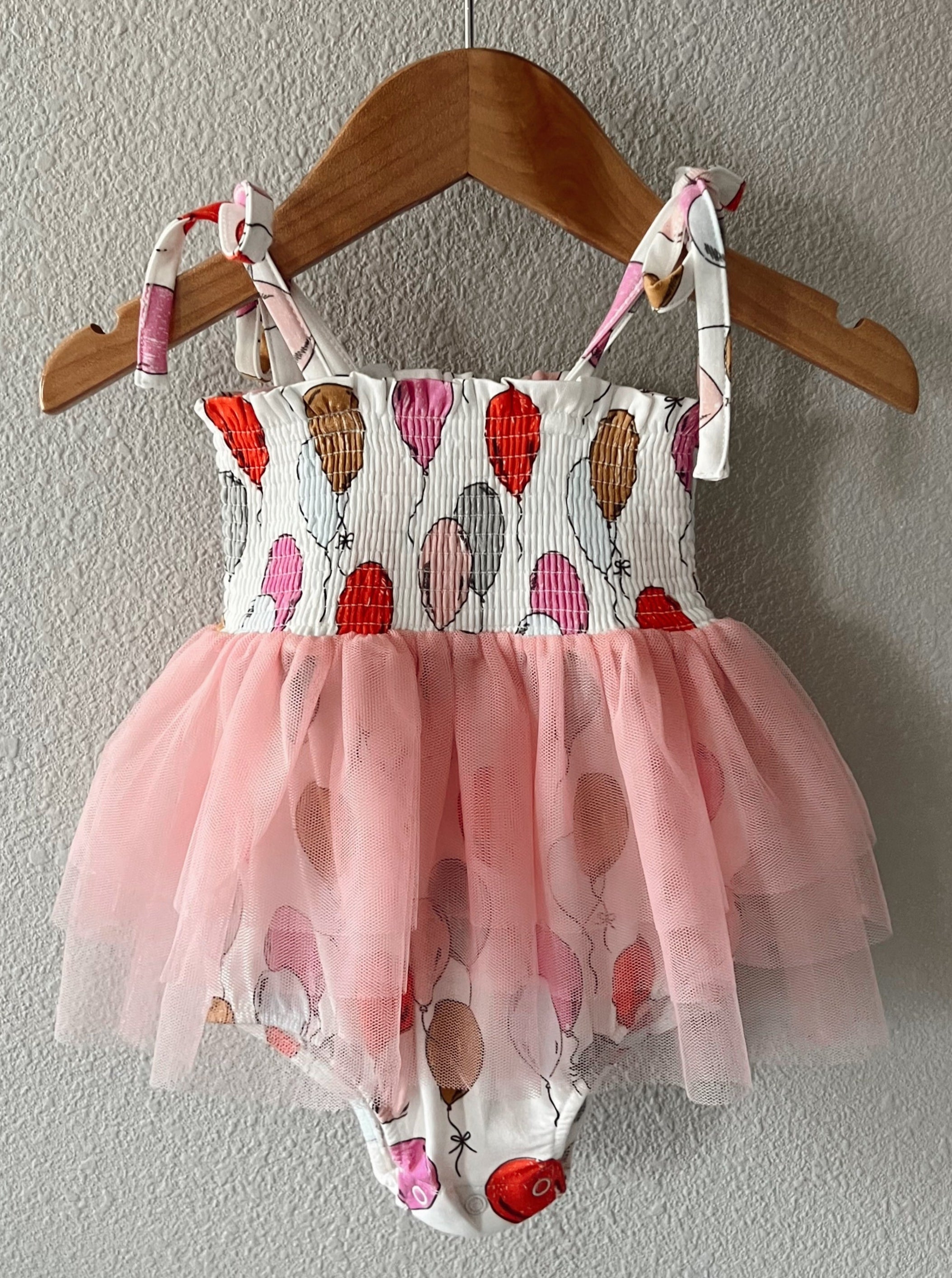 Child's dress featuring a balloon print with a pink tulle skirt, hanging on a wooden hanger against a textured wall.
