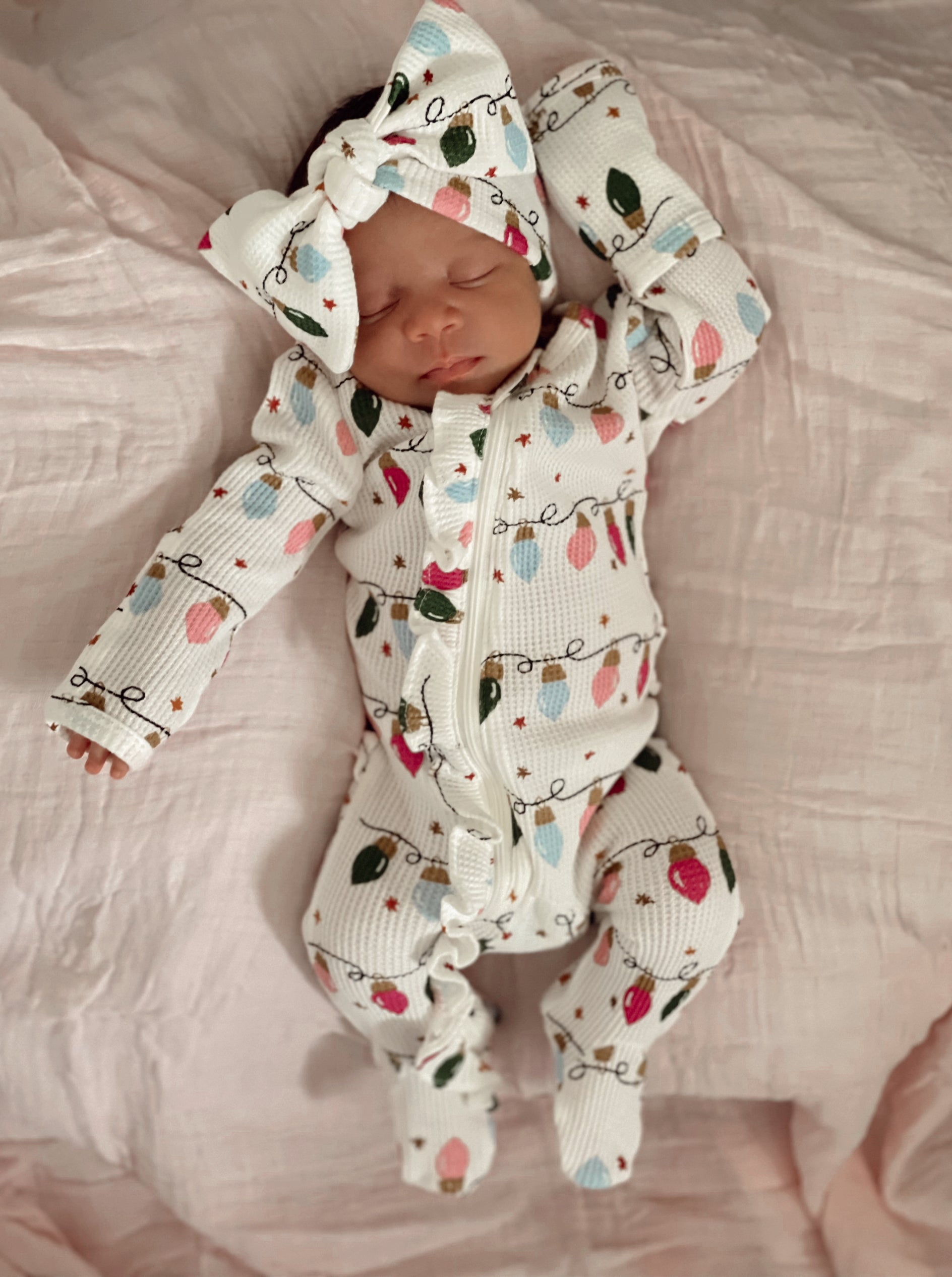Sleeping baby in a festive onesie with colorful lights and a large bow on a soft pink blanket.