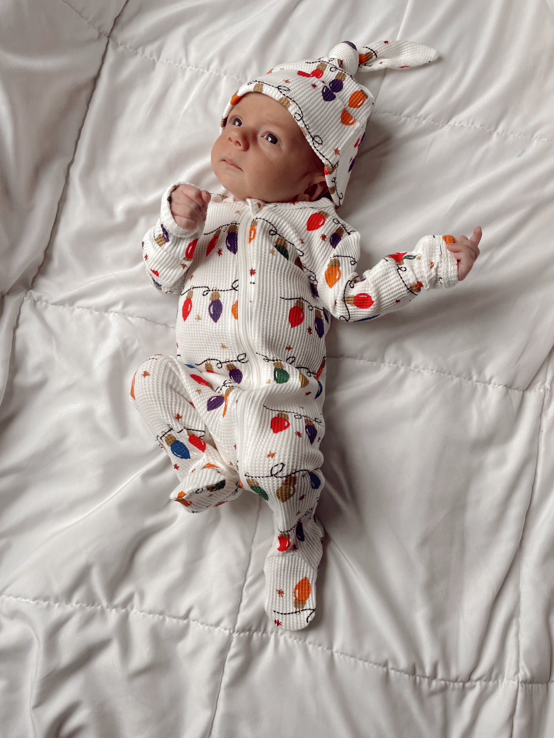 Baby in festive outfit with colorful lights pattern, lying on a white blanket, looking curiously at the camera.