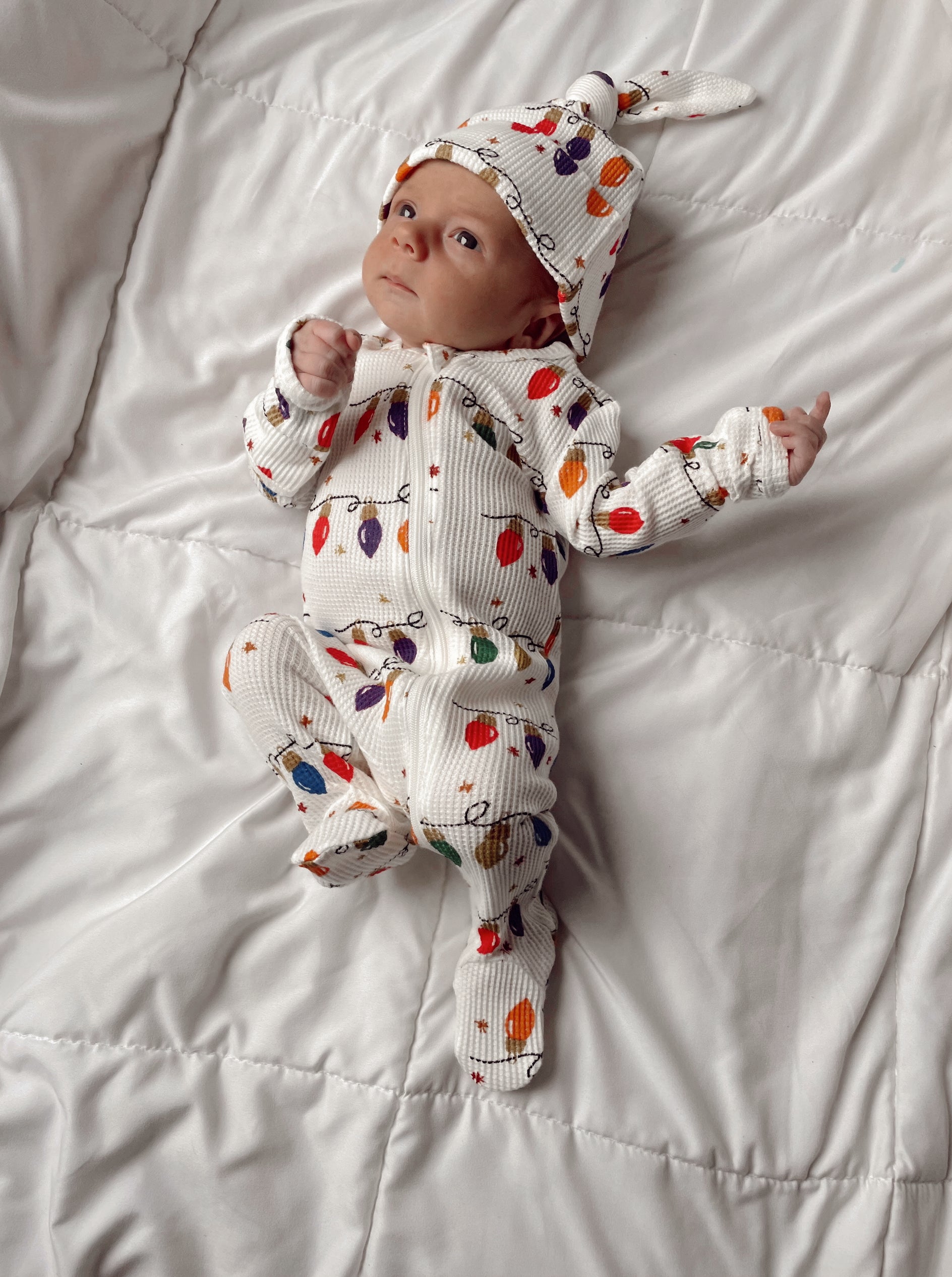 Baby in festive outfit with colorful lights pattern, lying on a white blanket, looking curiously at the camera.