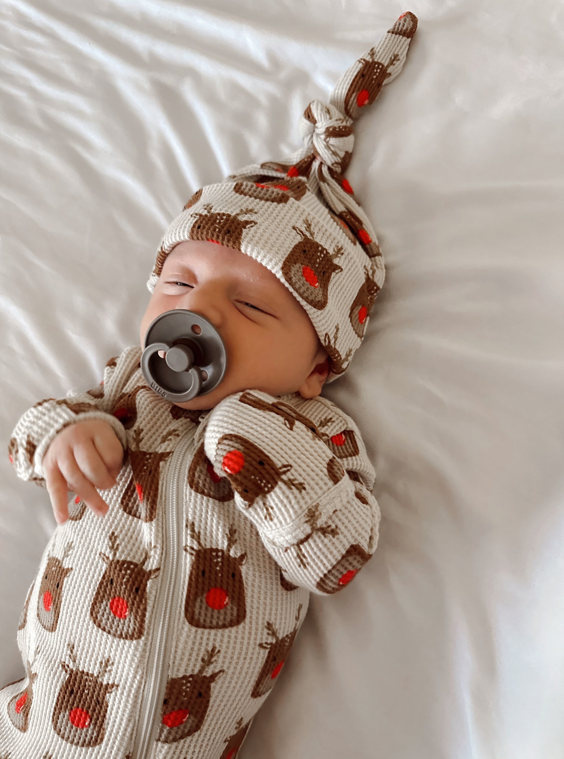 Baby wearing a reindeer-patterned outfit and hat, peacefully sleeping with a pacifier on a white bed.