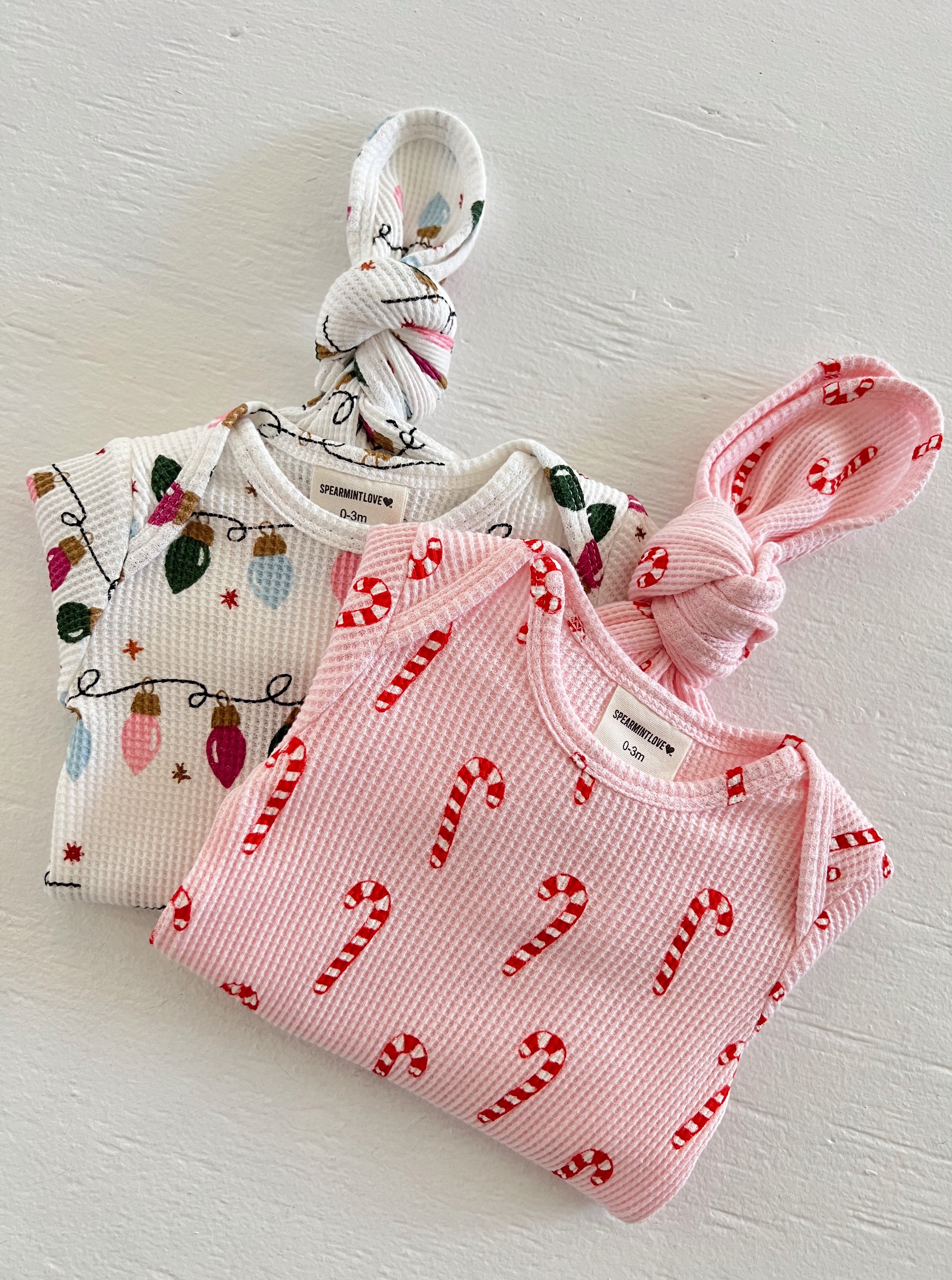 Two baby outfits: one with holiday lights and another pink with candy cane prints, both featuring knotted tops.