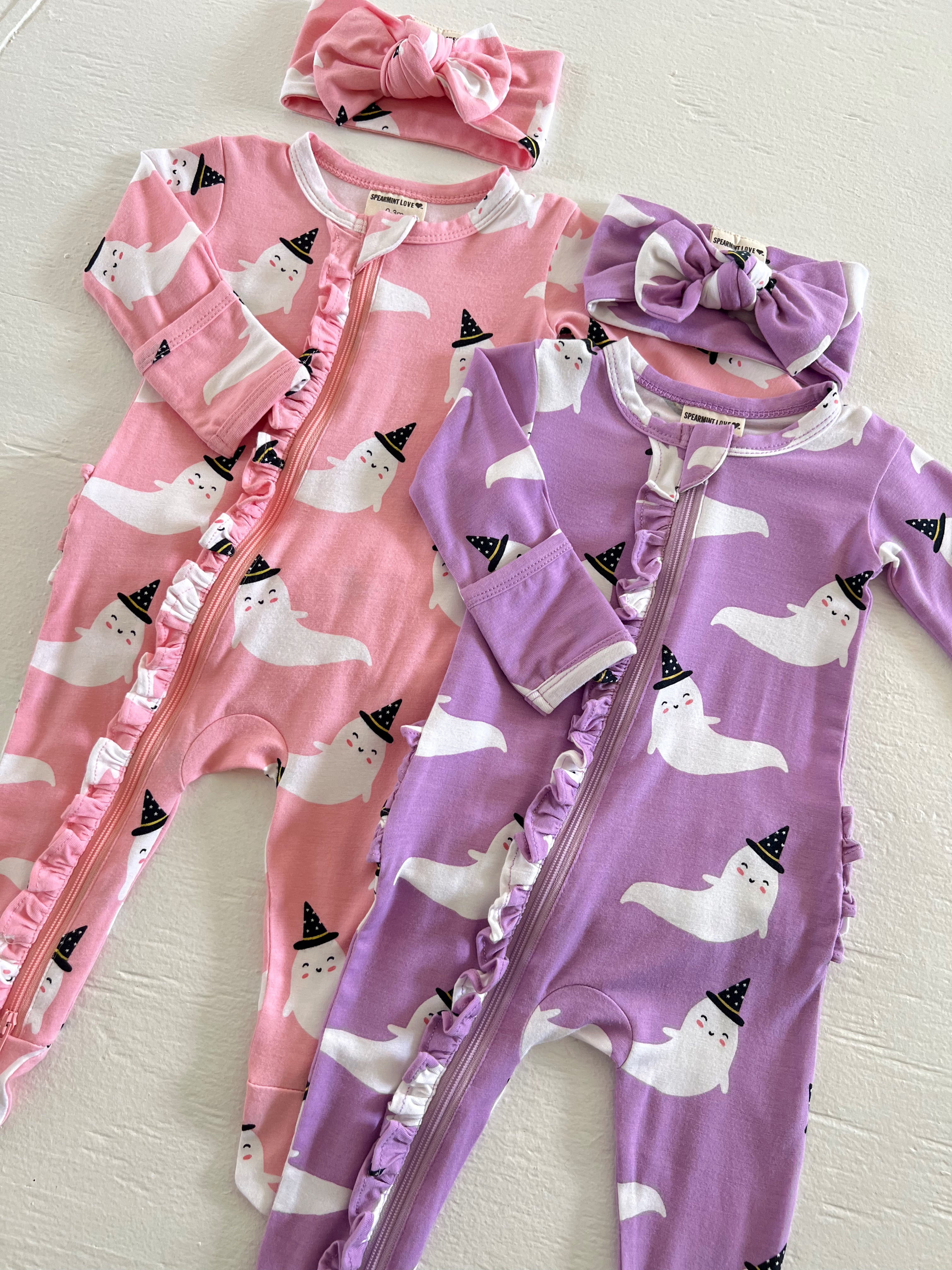 Two ghost-themed baby onesies in pink and purple with matching headbands, featuring ruffles and party hats.