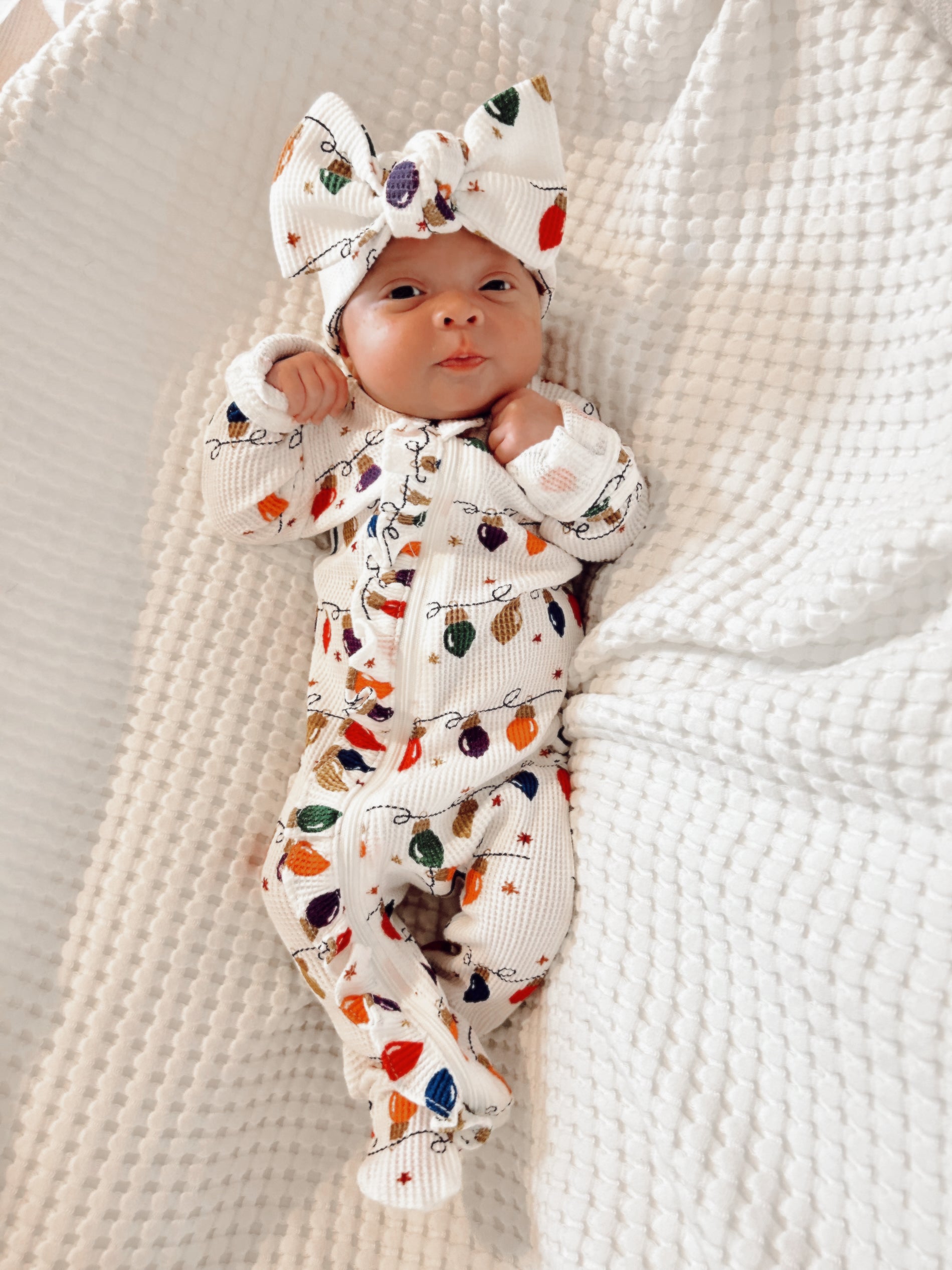 Baby in colorful holiday-themed outfit with a large bow, lying on a textured white blanket.
