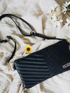 Black crossbody bag on a textured fabric background, with a small sunflower nearby and white flowers in the scene.