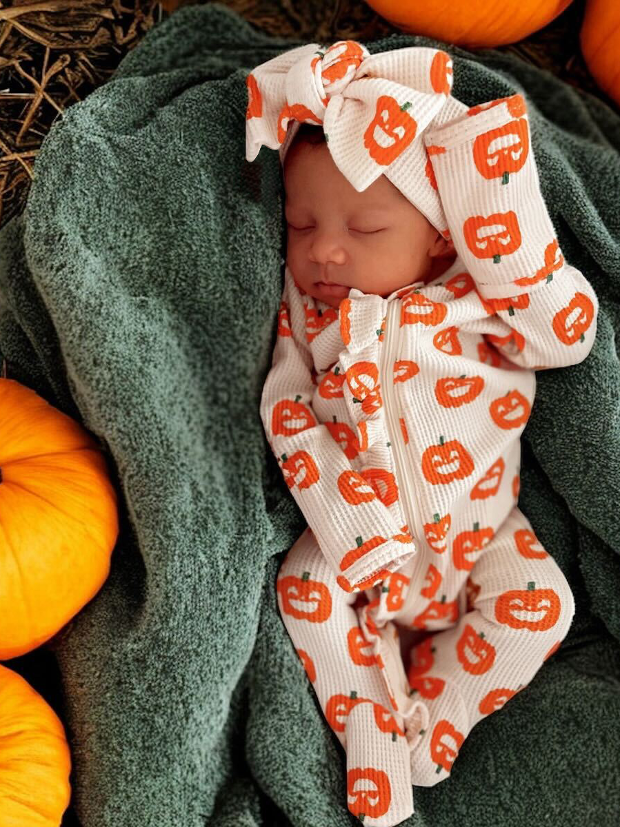 Sleeping baby in pumpkin-patterned outfit, wrapped in a green blanket, surrounded by orange pumpkins.