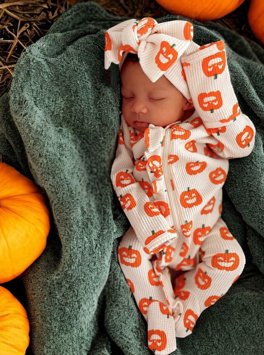 Sleeping baby in pumpkin-patterned outfit, wrapped in a green blanket, surrounded by orange pumpkins.