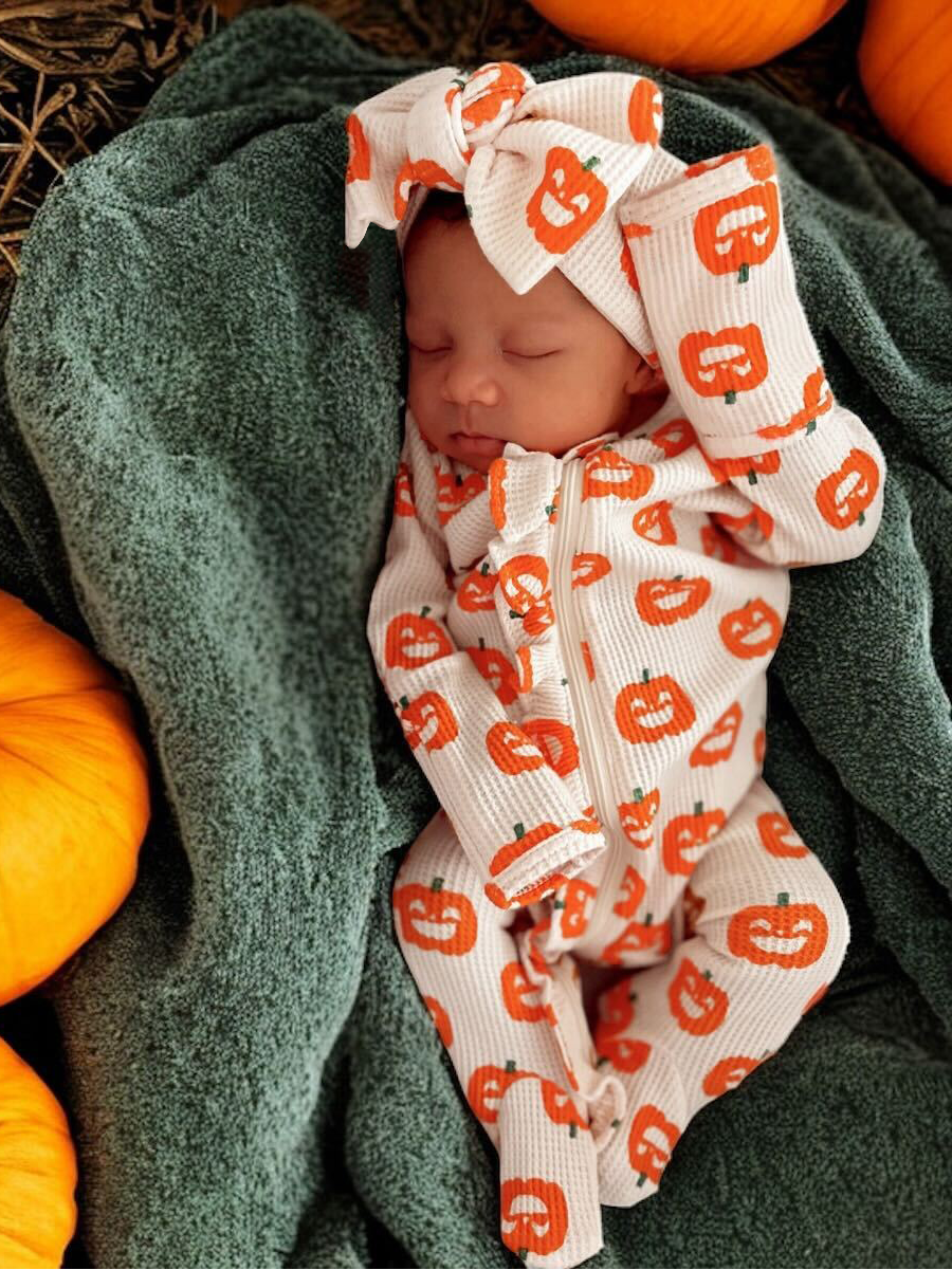 Sleeping baby in pumpkin-patterned outfit, wrapped in a green blanket, surrounded by orange pumpkins.