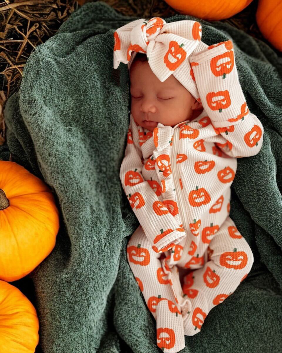 Sleeping baby in pumpkin-patterned outfit, wrapped in a green blanket, surrounded by orange pumpkins.