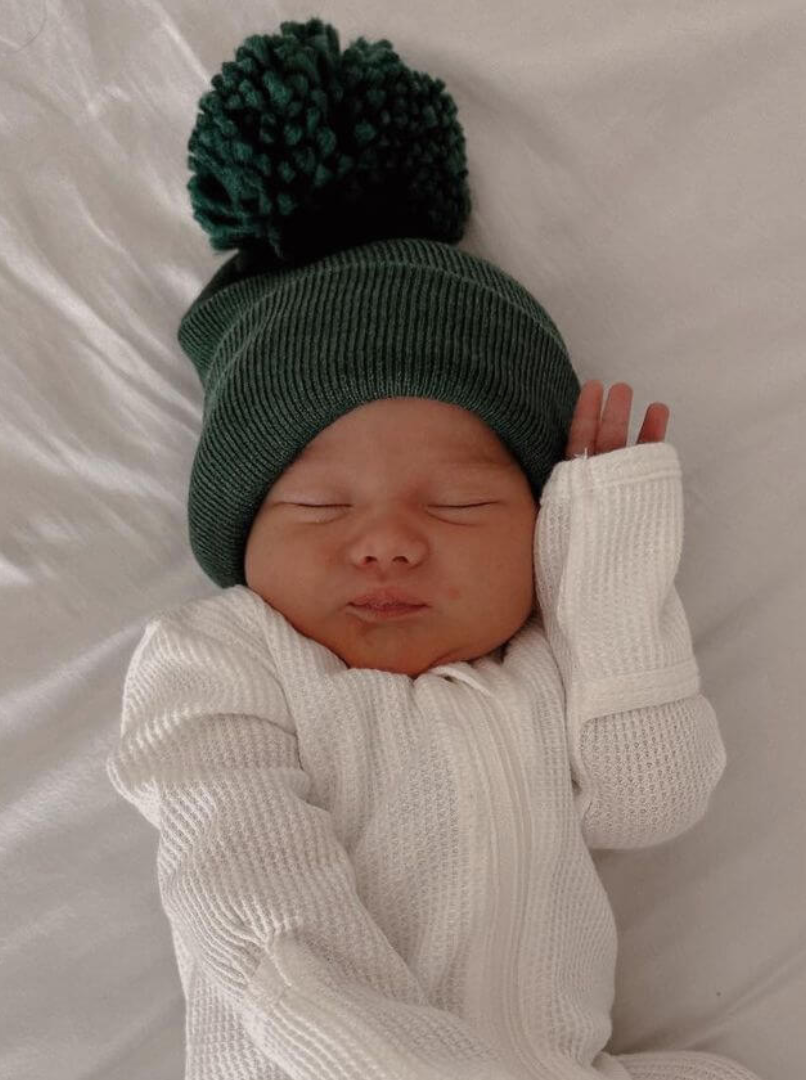 Newborn baby sleeping peacefully in a cozy green hat and white outfit on a soft white surface.