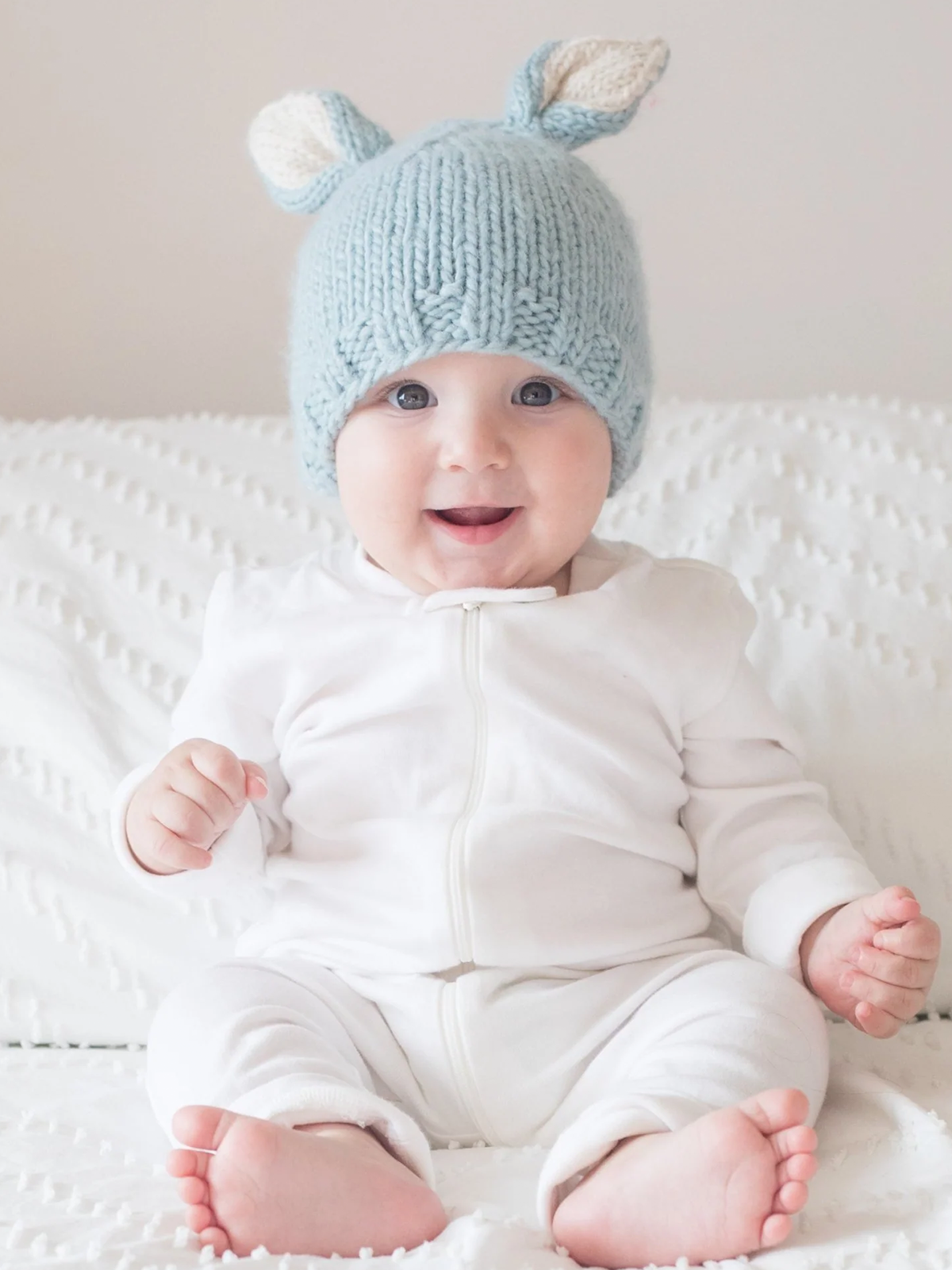 Smiling baby in a light blue knitted hat with ears, wearing a white outfit, sitting on a textured blanket.