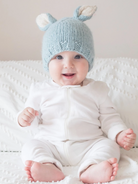 Smiling baby in a light blue knitted hat with ears, wearing a white outfit, sitting on a textured blanket.