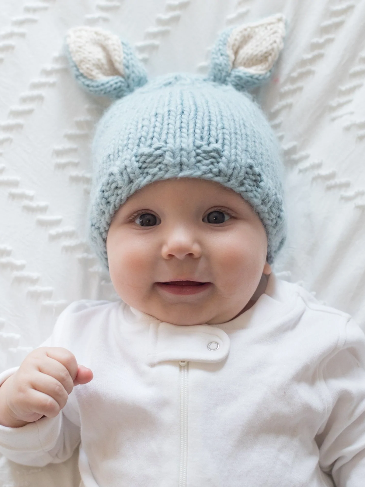 Smiling baby wearing a light blue knitted hat with bunny ears, lying on a textured white blanket.