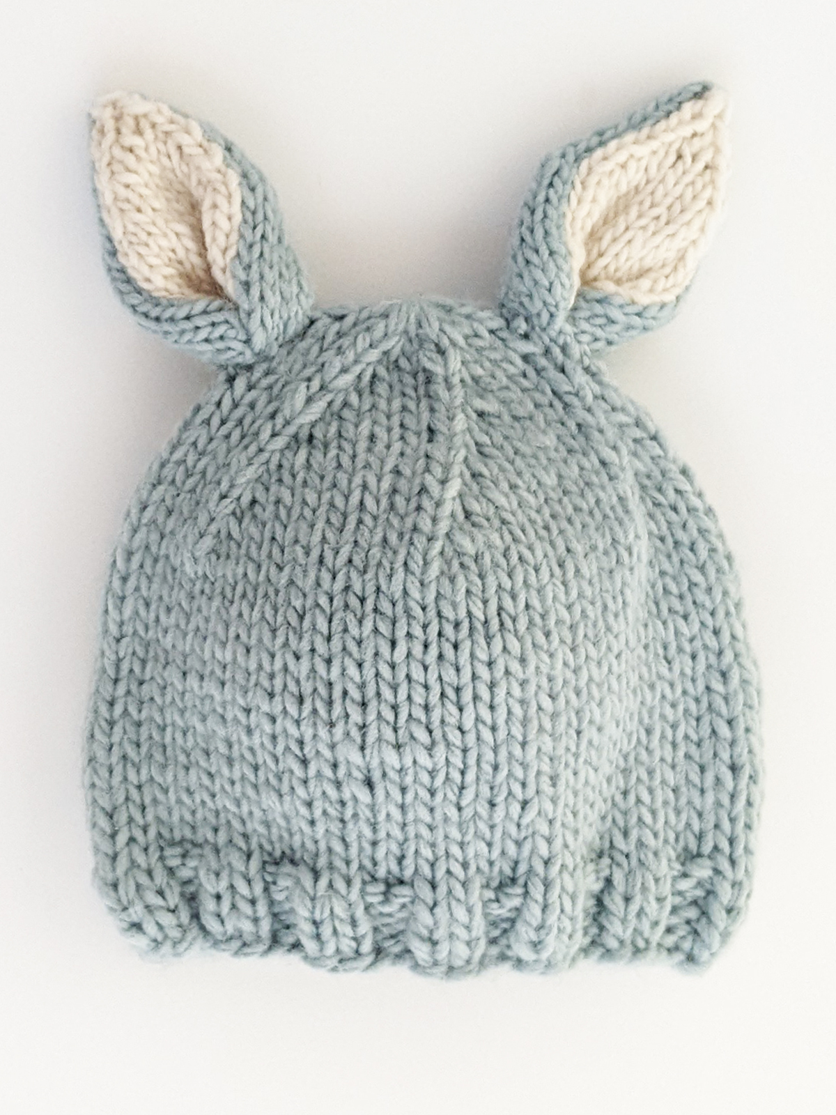 Light blue knitted hat with cream bunny ears on top, resting on a white background.