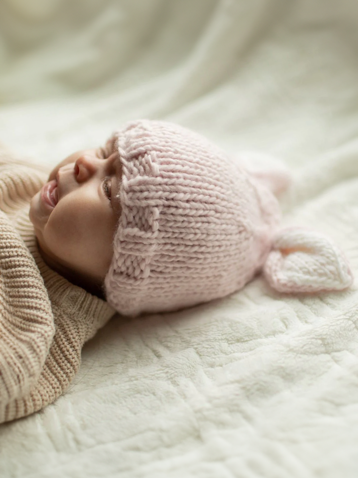 Baby in a pink knitted hat with ears, lying on a soft blanket, making a playful expression.