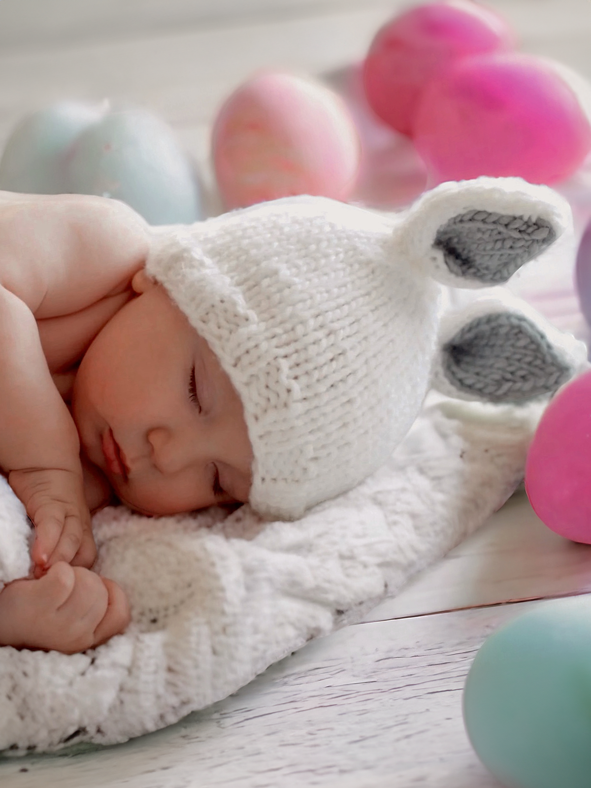 Sleeping baby in a white bunny hat surrounded by colorful Easter eggs on a knitted blanket.