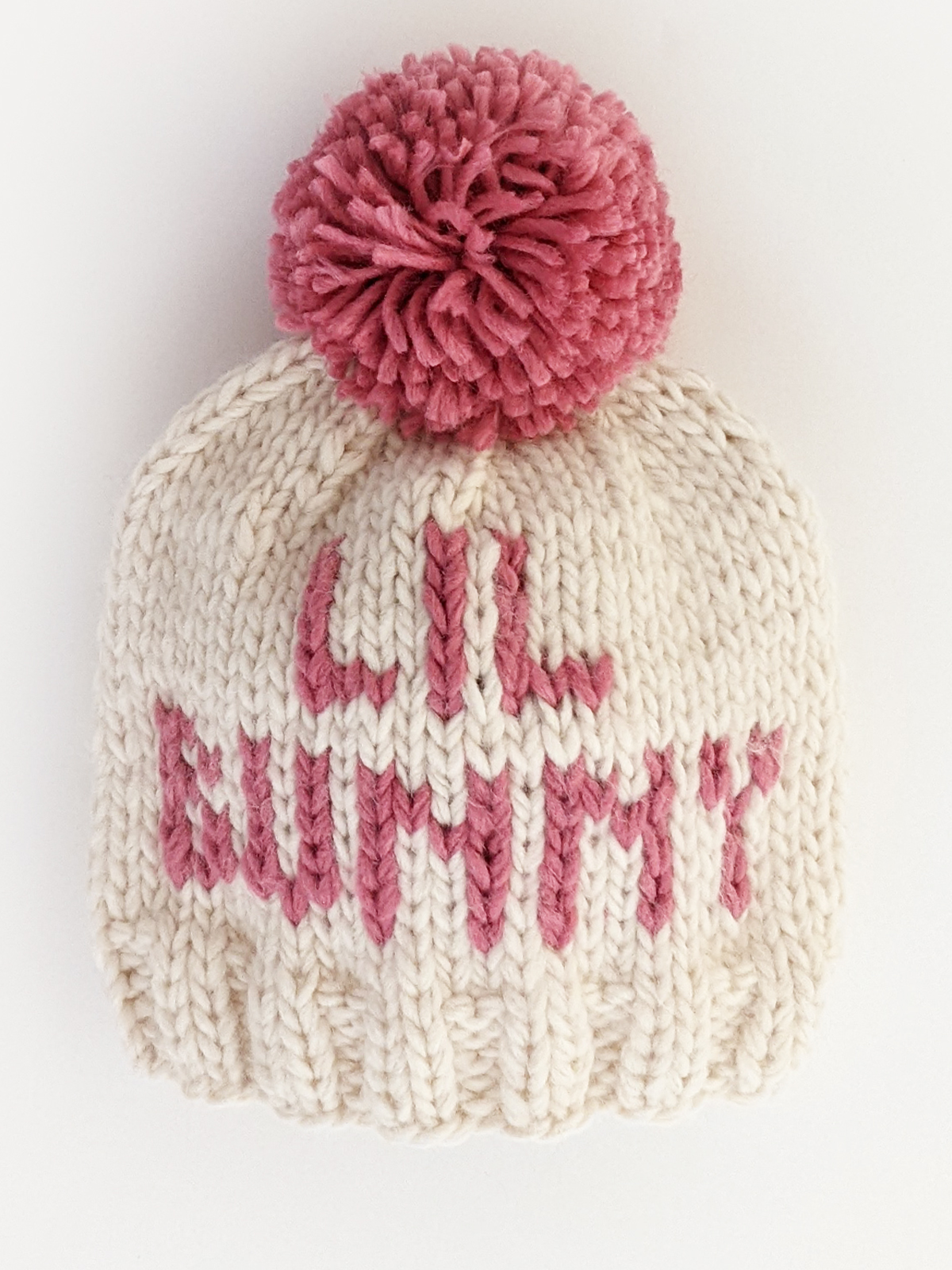 Cream-colored knitted baby hat with pink pom-pom and "Lil Bunny" text.