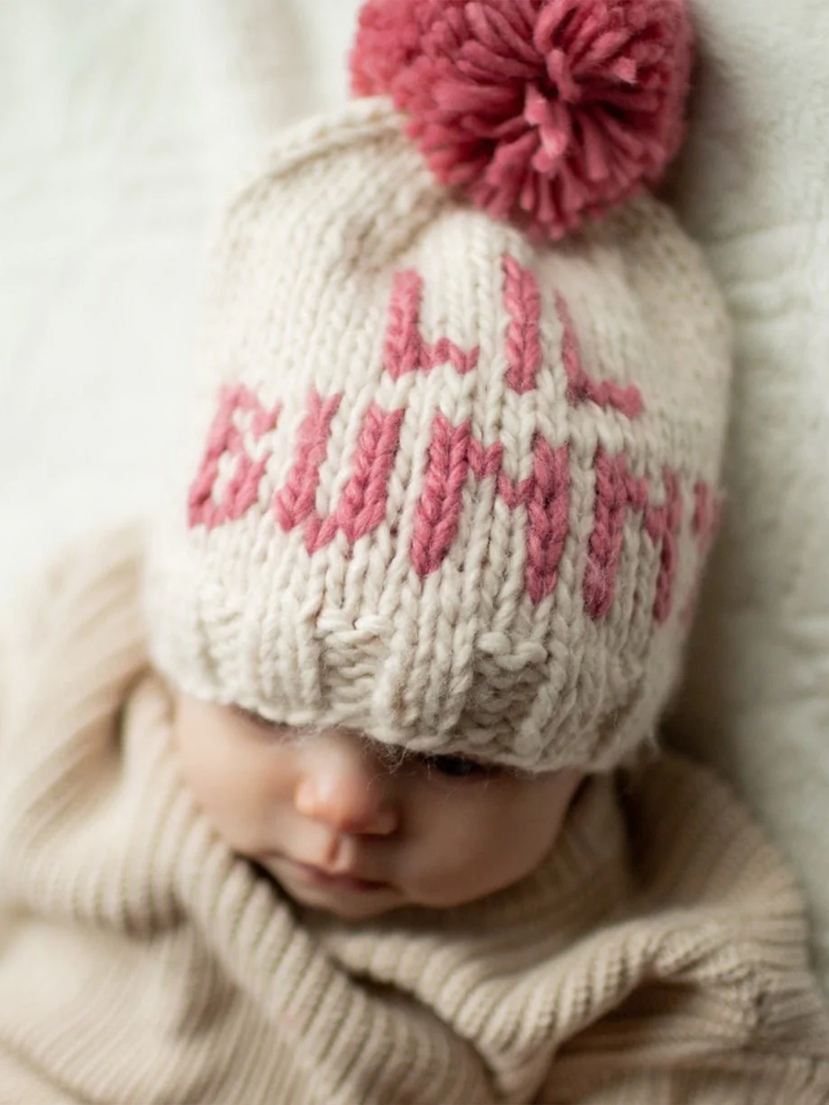 Baby wearing a knitted cream hat with pink pom-pom and the words "Hi Bunny," looking down.
