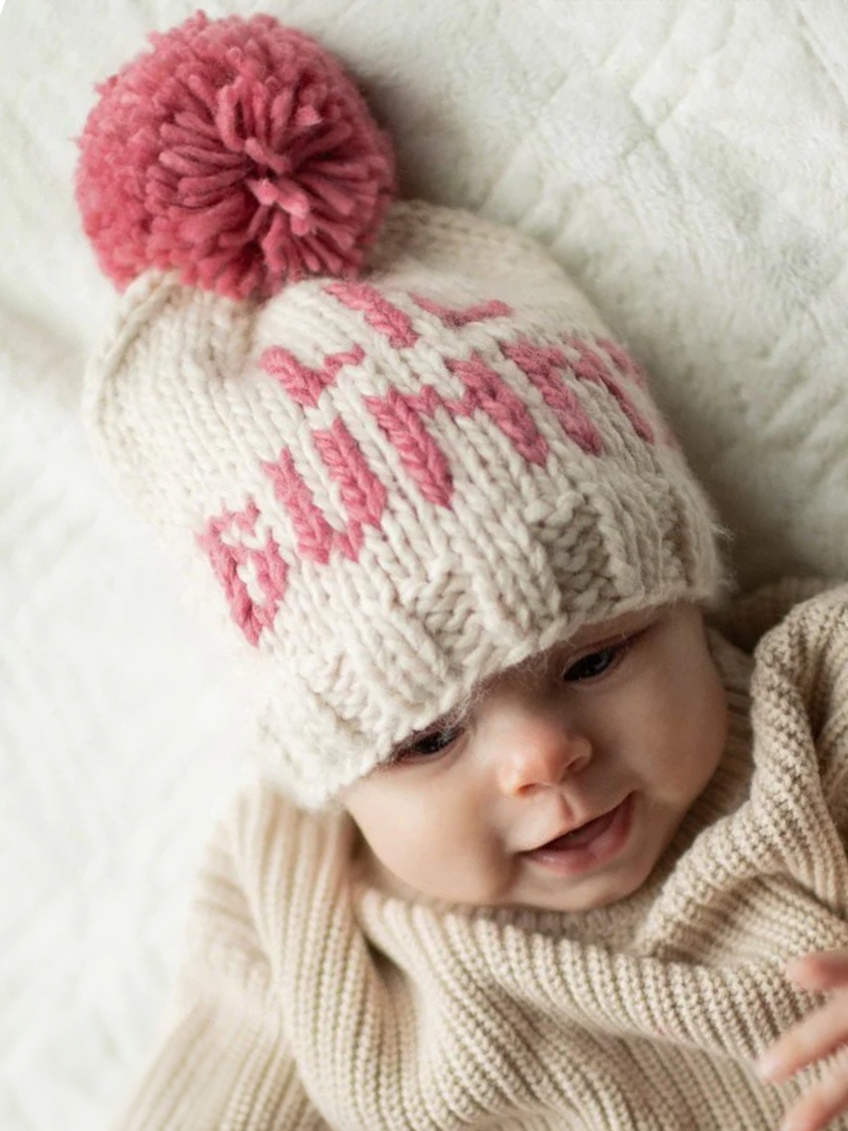Smiling baby in a cozy sweater wearing a knitted hat with pink pom-pom and playful lettering.