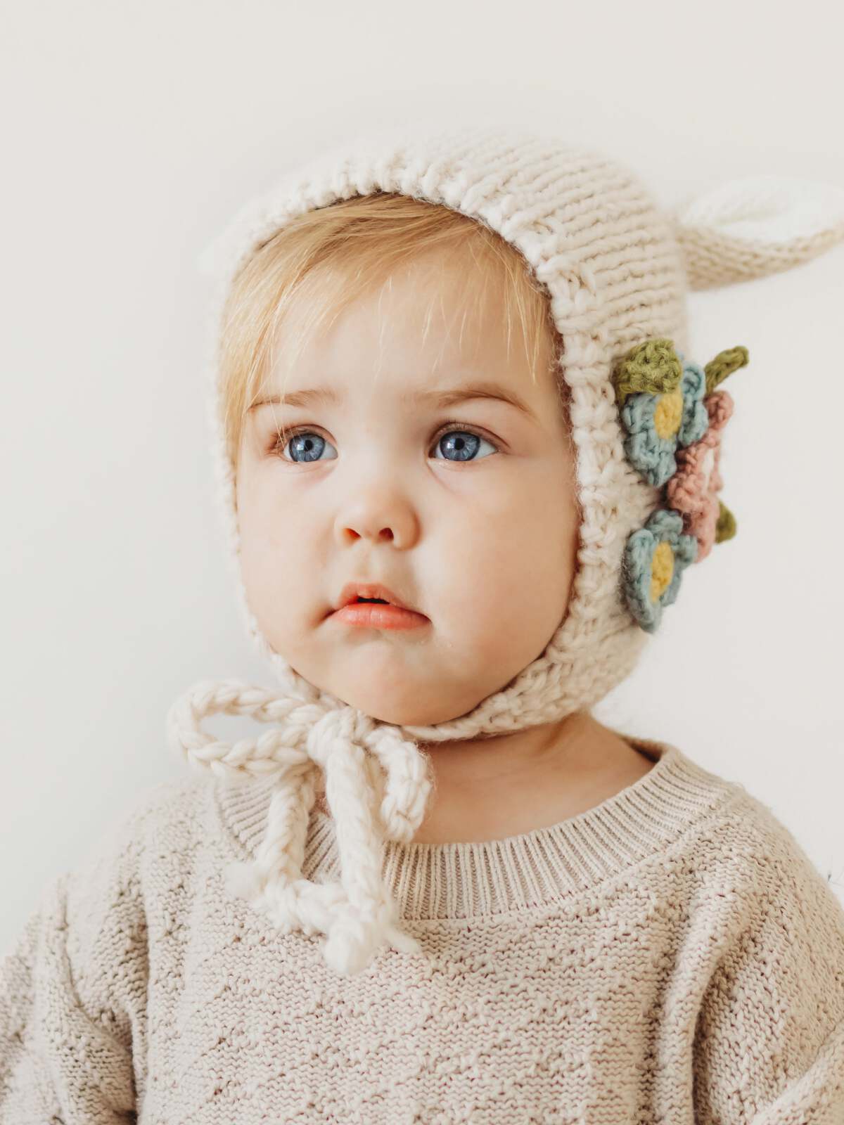 Toddler wearing a knitted hat with a flower design, looking thoughtfully against a plain background.