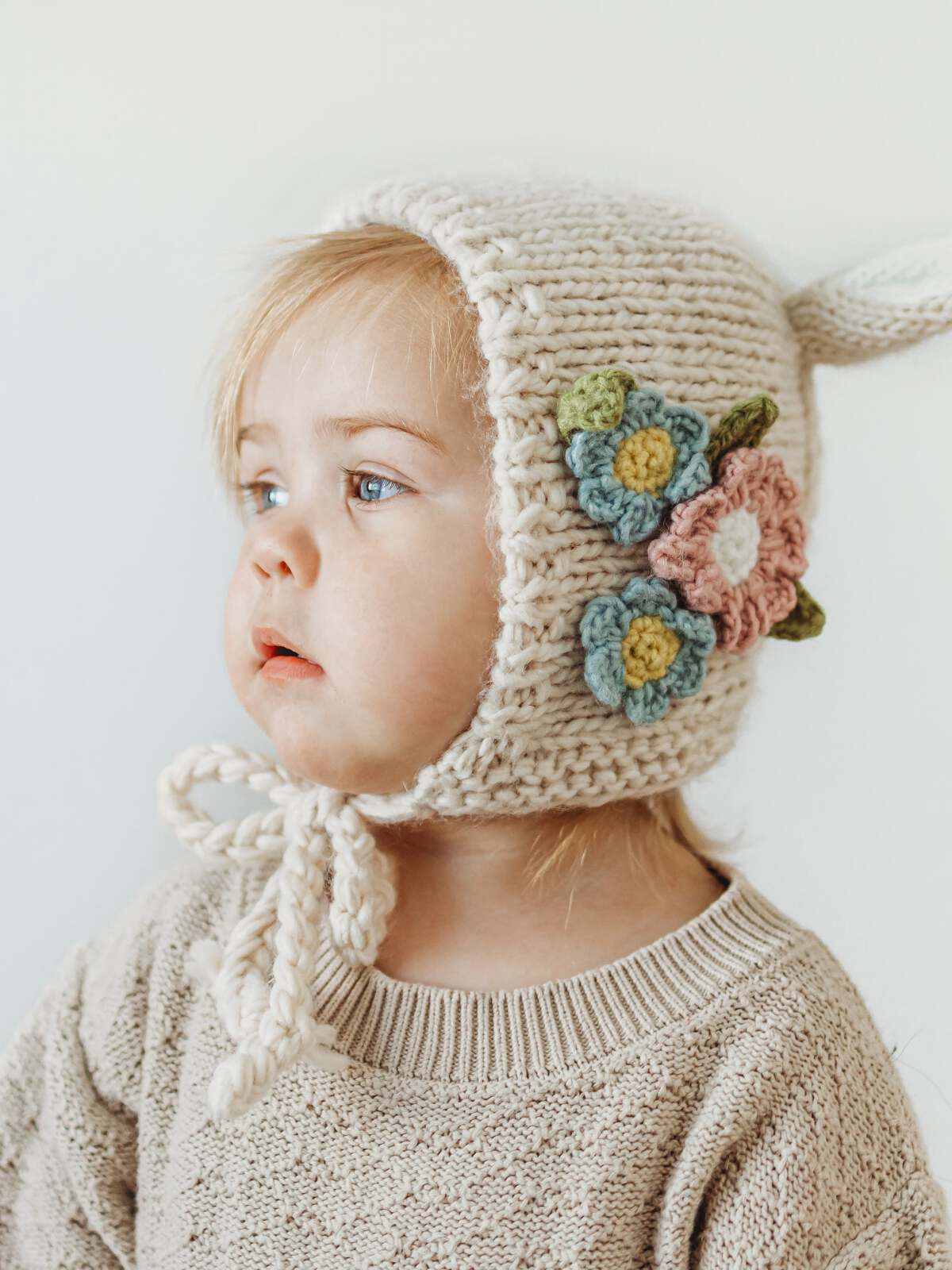Toddler in a knitted floral bonnet, gazing thoughtfully, wearing a cozy sweater.
