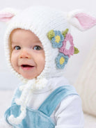 Baby smiling while wearing a cozy white knit hat with bunny ears and colorful flowers. Soft background.