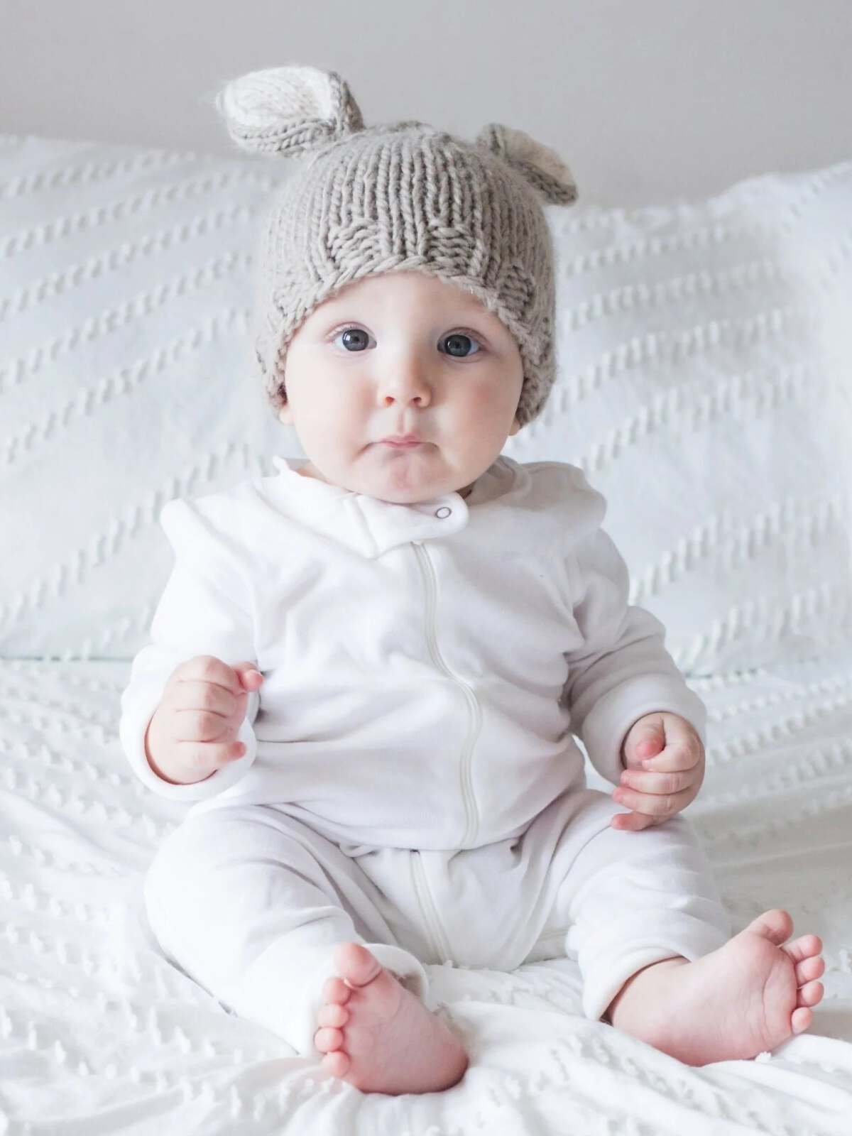 Baby in a fluffy gray bear hat sits on a white blanket, looking curiously at the camera.