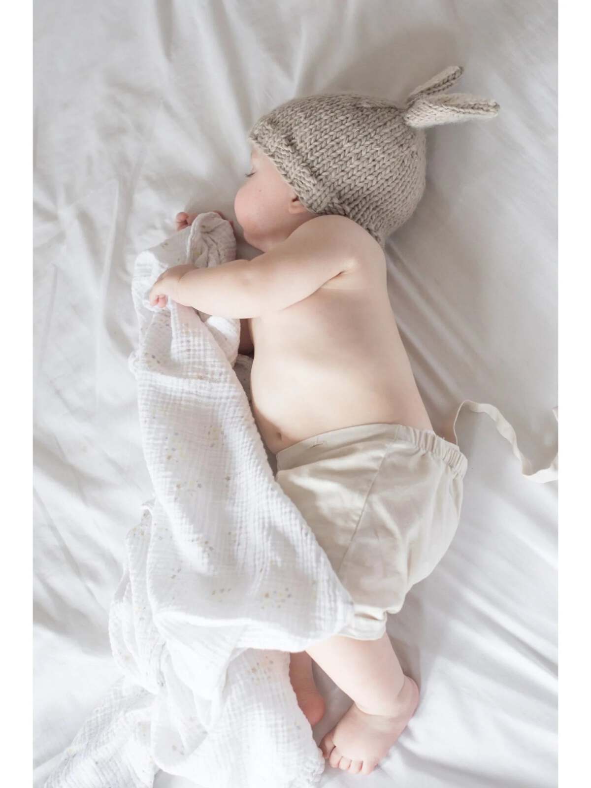Baby wearing a knitted bunny hat, sleeping peacefully with a soft blanket on a light-colored bed.