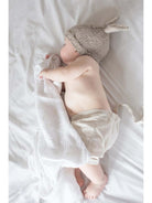 Baby wearing a knitted bunny hat, sleeping peacefully with a soft blanket on a light-colored bed.