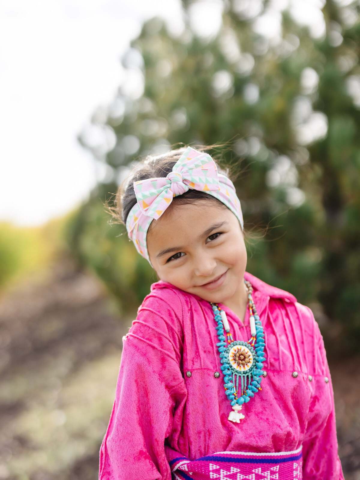 Smiling girl in a pink outfit with a patterned headband and necklace, standing outdoors with greenery behind her.