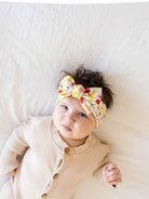 Baby with curly hair wearing a colorful headband, lying on a soft white surface with a serene expression.