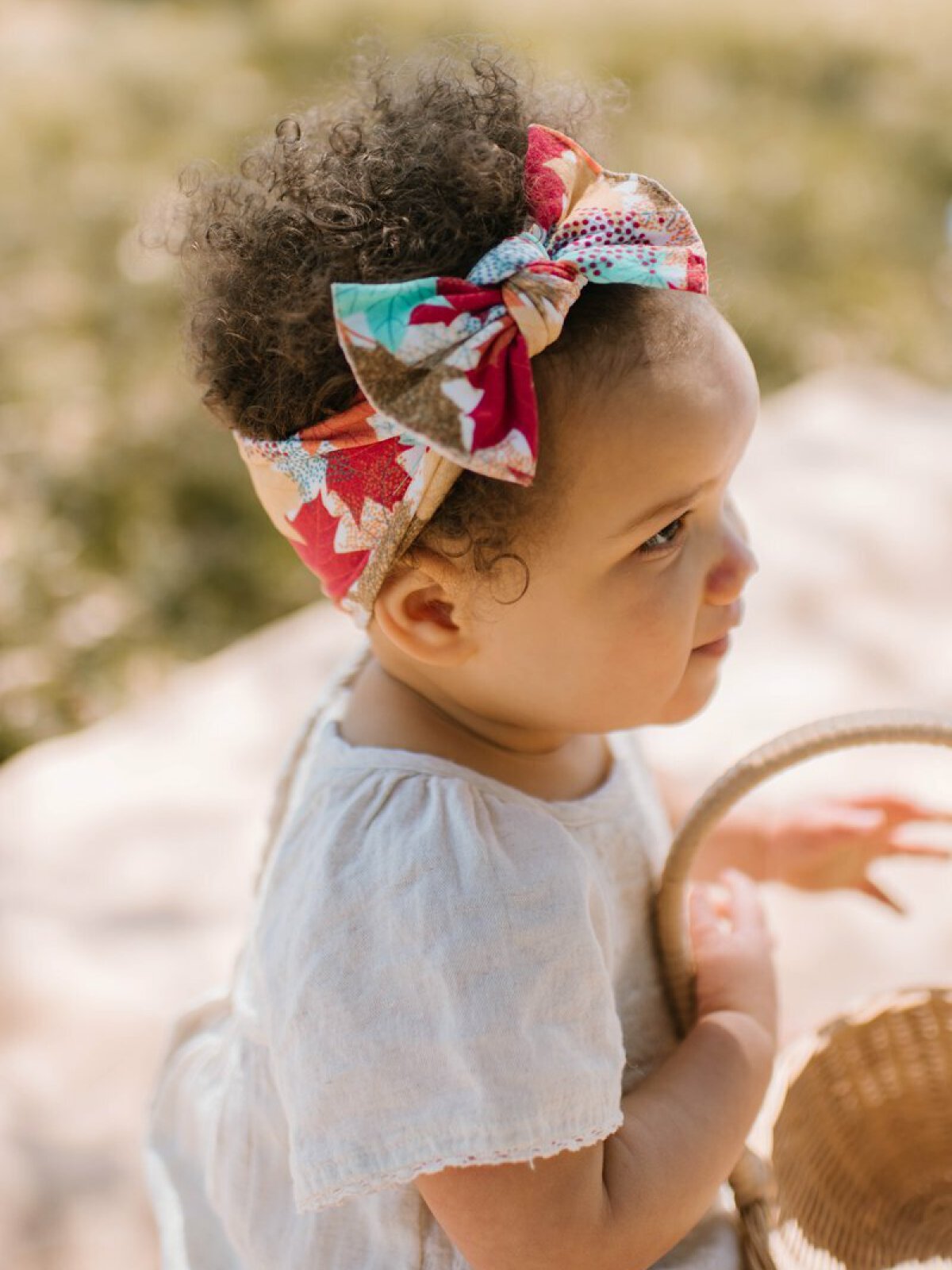 Toddler with curly hair wearing a colorful bow, holding a small woven basket outdoors.