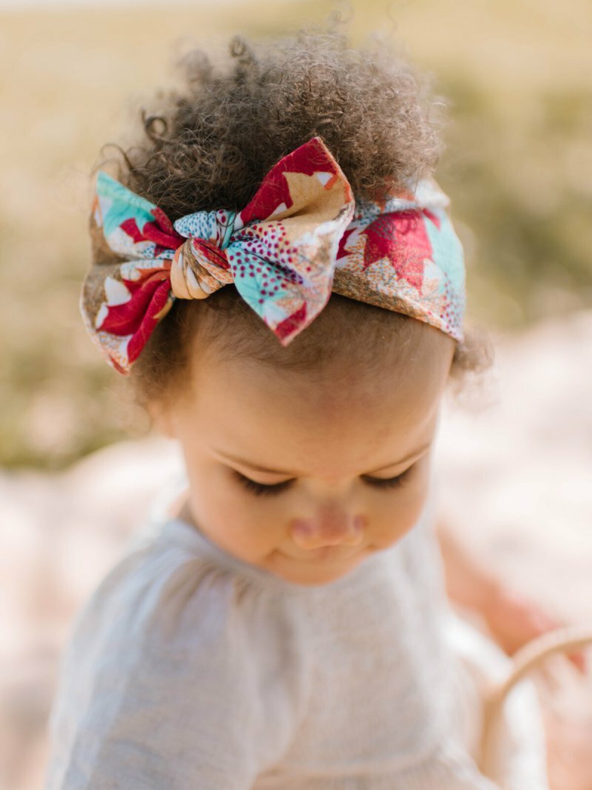 Toddler with curly hair wearing a colorful patterned headband, looking down while outdoors.