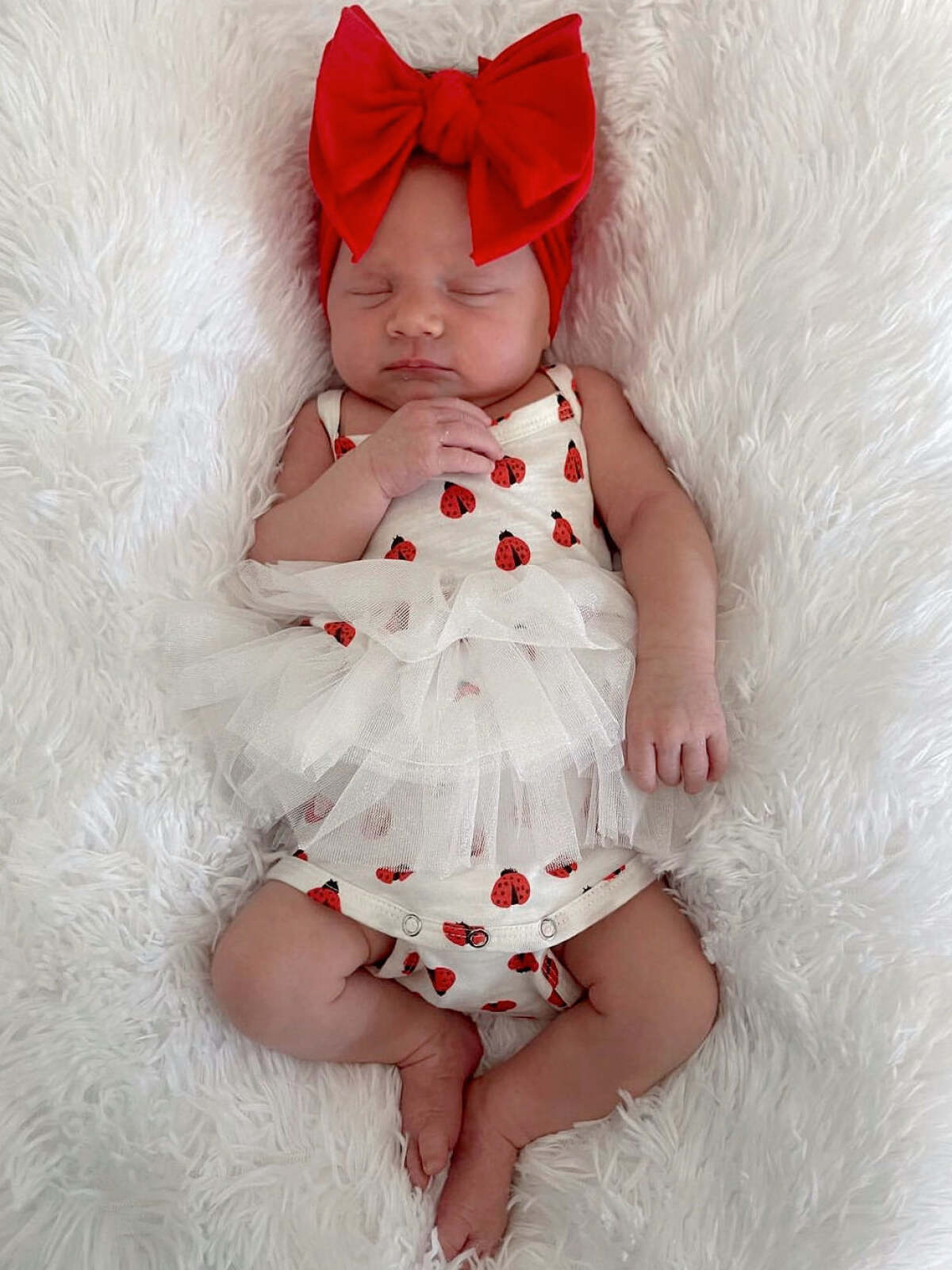 Baby girl sleeping on a fluffy white surface, wearing a ladybug onesie and a large red bow headband.