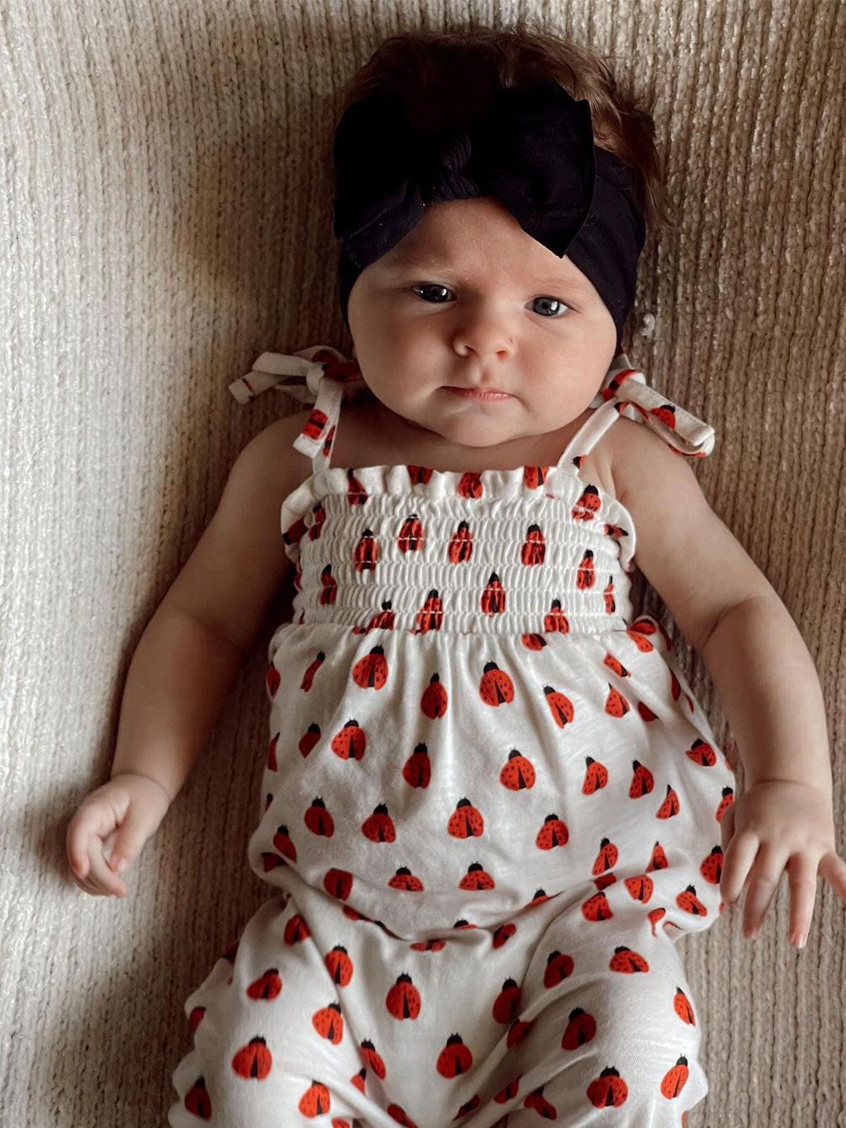 Infant girl lying on a textured surface, wearing a white outfit with red ladybug patterns and a black headband.