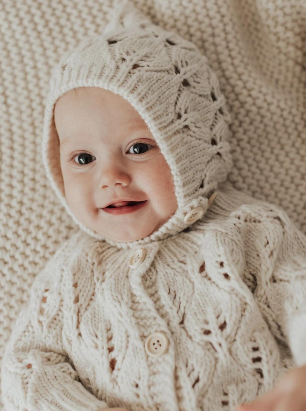 Smiling baby in a cozy cream knitted outfit and hat, resting on a matching blanket.
