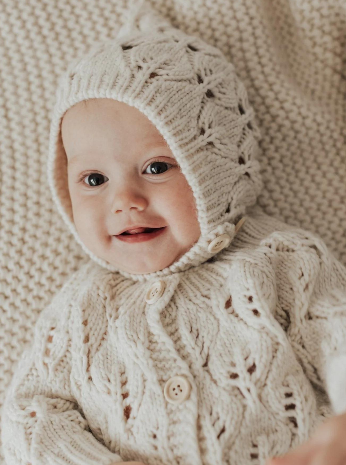 Smiling baby in a cozy cream knitted outfit and hat, resting on a matching blanket.