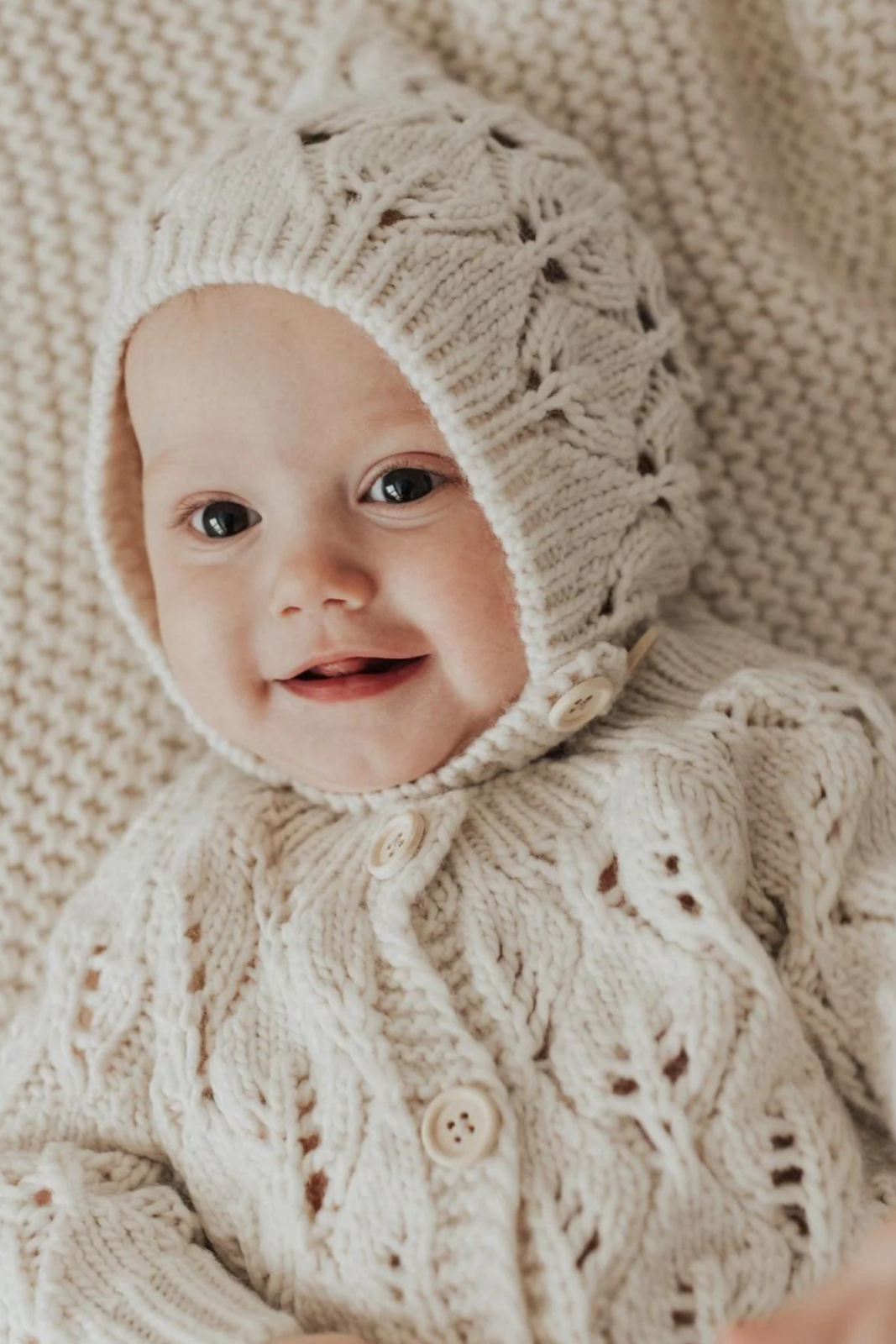 Smiling baby in a cozy cream knitted outfit and hat, resting on a matching blanket.