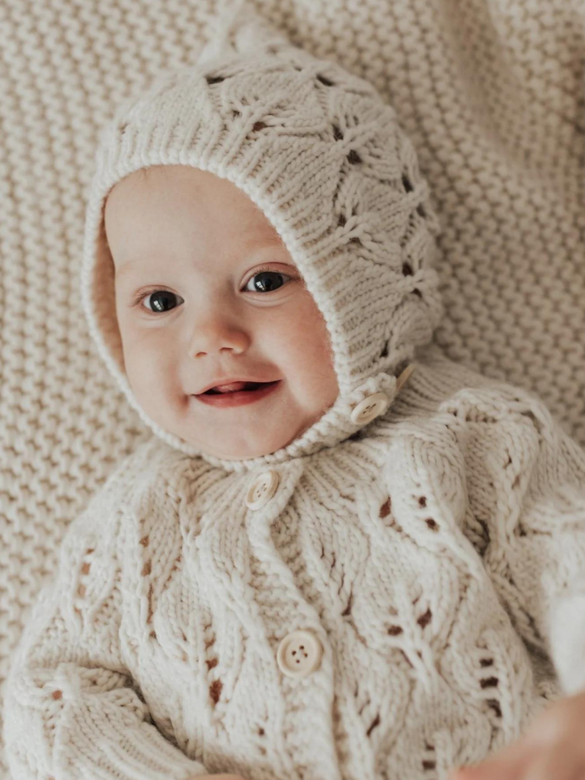 Smiling baby in a cozy cream knitted outfit and hat, resting on a matching blanket.