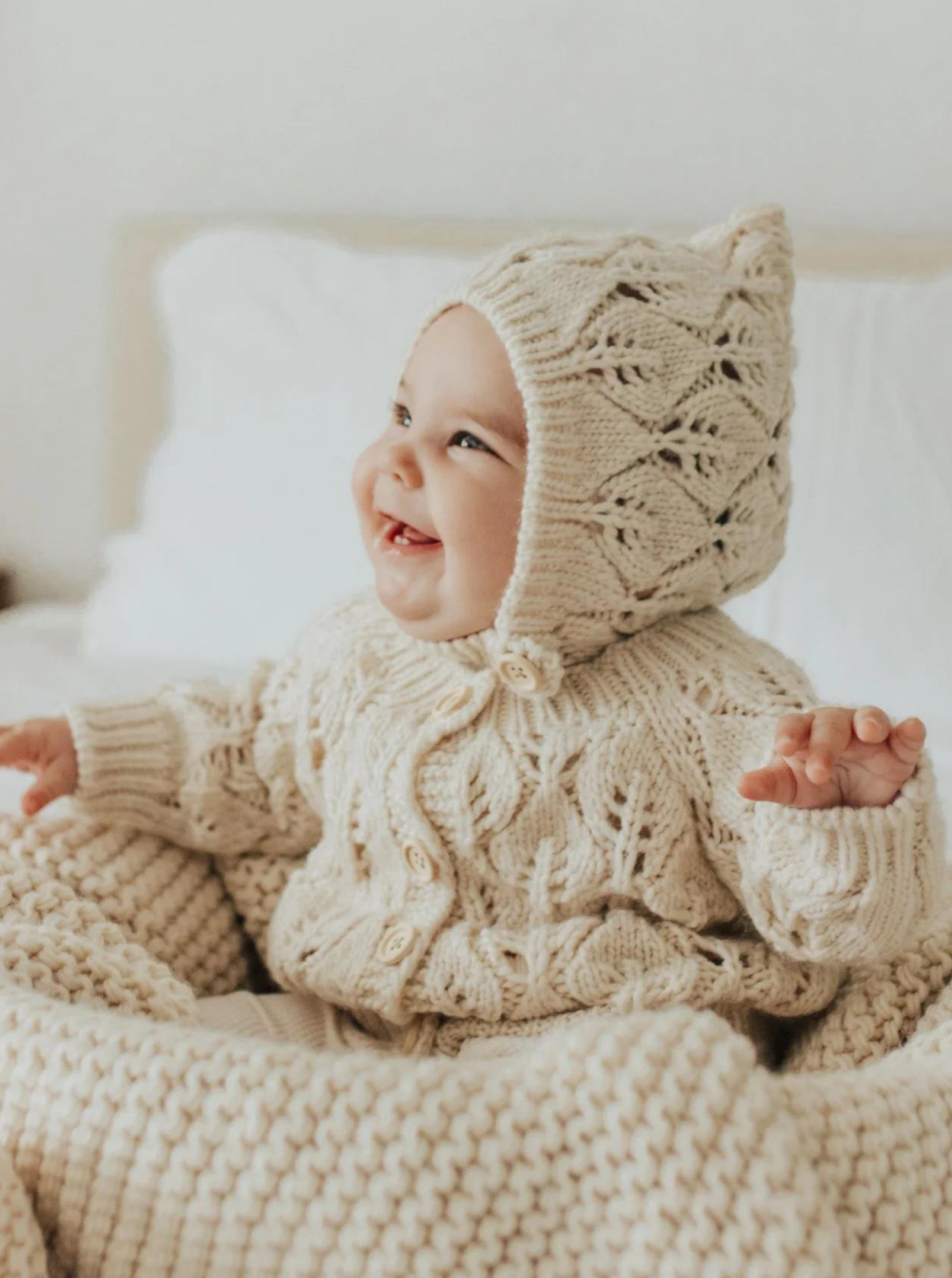 Smiling baby in cozy, knitted cream outfit and bonnet, sitting on a soft blanket in a bright, airy room.
