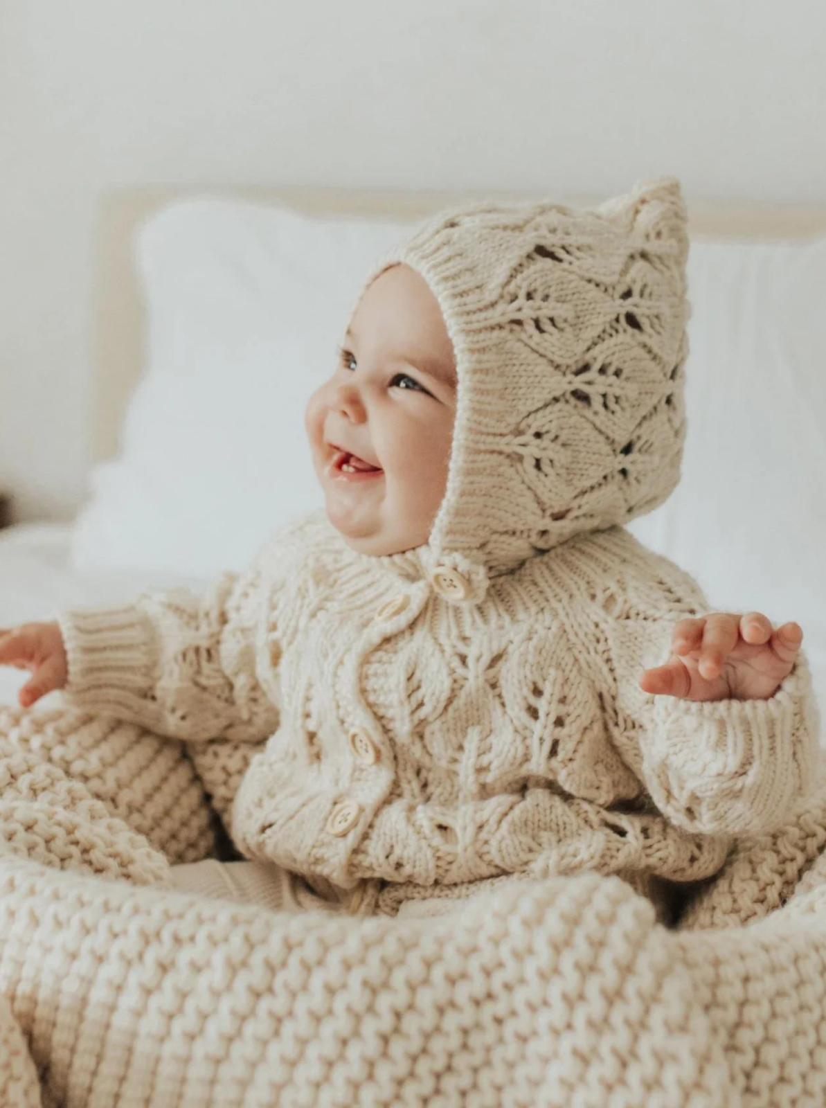 Smiling baby in cozy, knitted cream outfit and bonnet, sitting on a soft blanket in a bright, airy room.