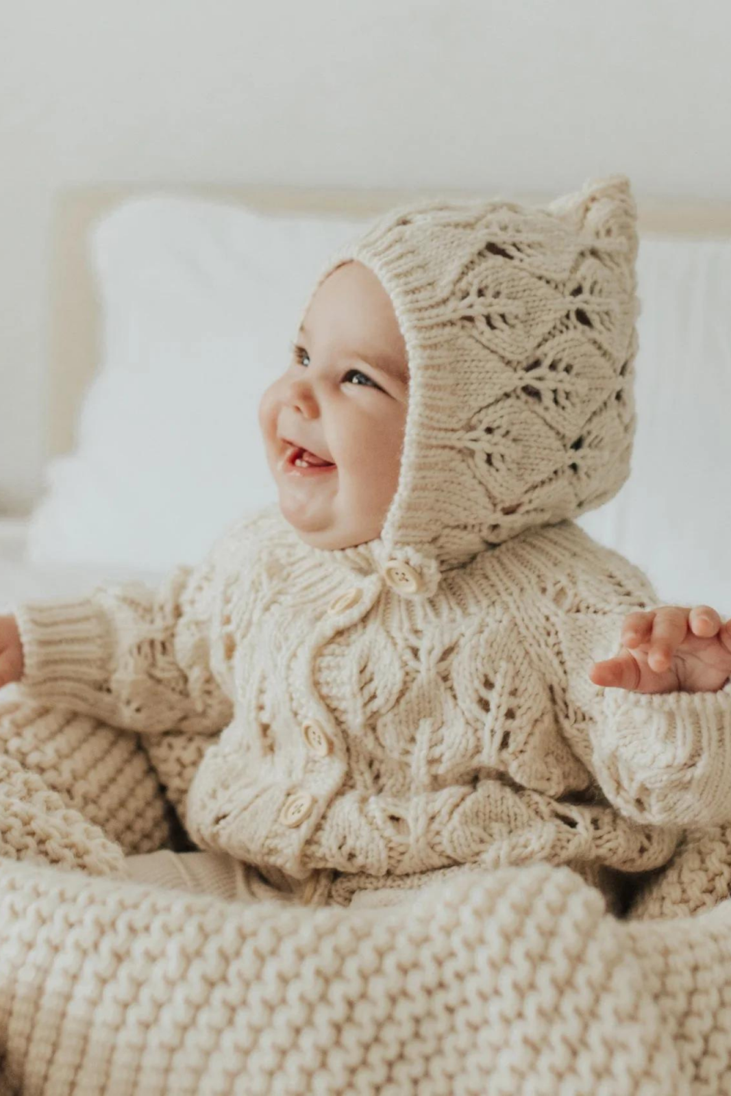 Smiling baby in cozy, knitted cream outfit and bonnet, sitting on a soft blanket in a bright, airy room.