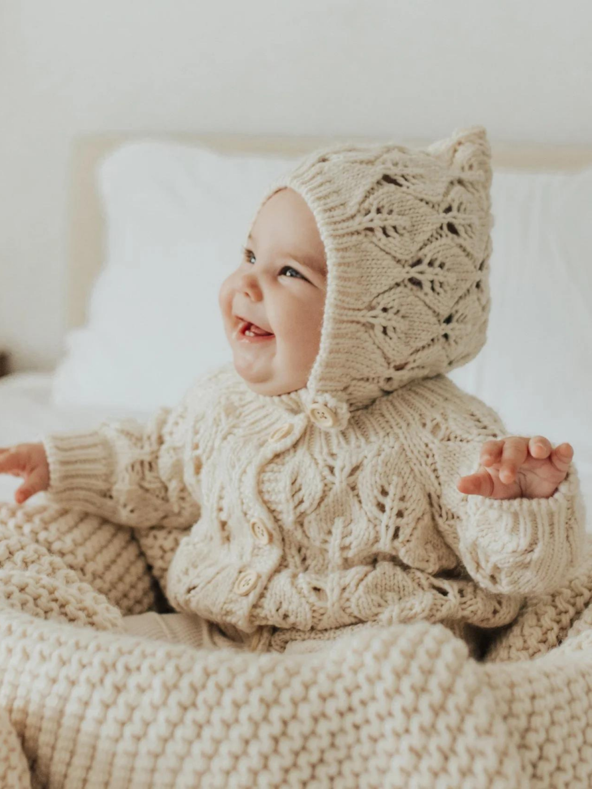 Smiling baby in cozy, knitted cream outfit and bonnet, sitting on a soft blanket in a bright, airy room.