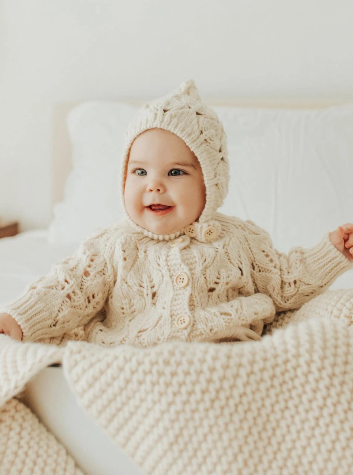 Smiling baby in a cozy, cream-colored knitted sweater and hat, sitting on a bed with blankets.