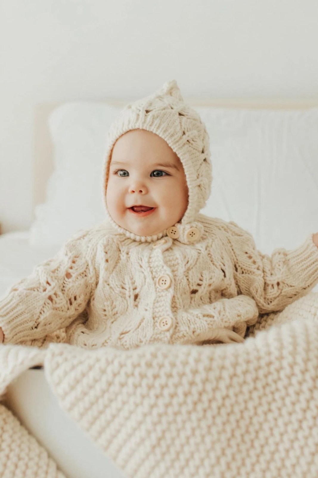 Smiling baby in a cozy, cream-colored knitted sweater and hat, sitting on a bed with blankets.